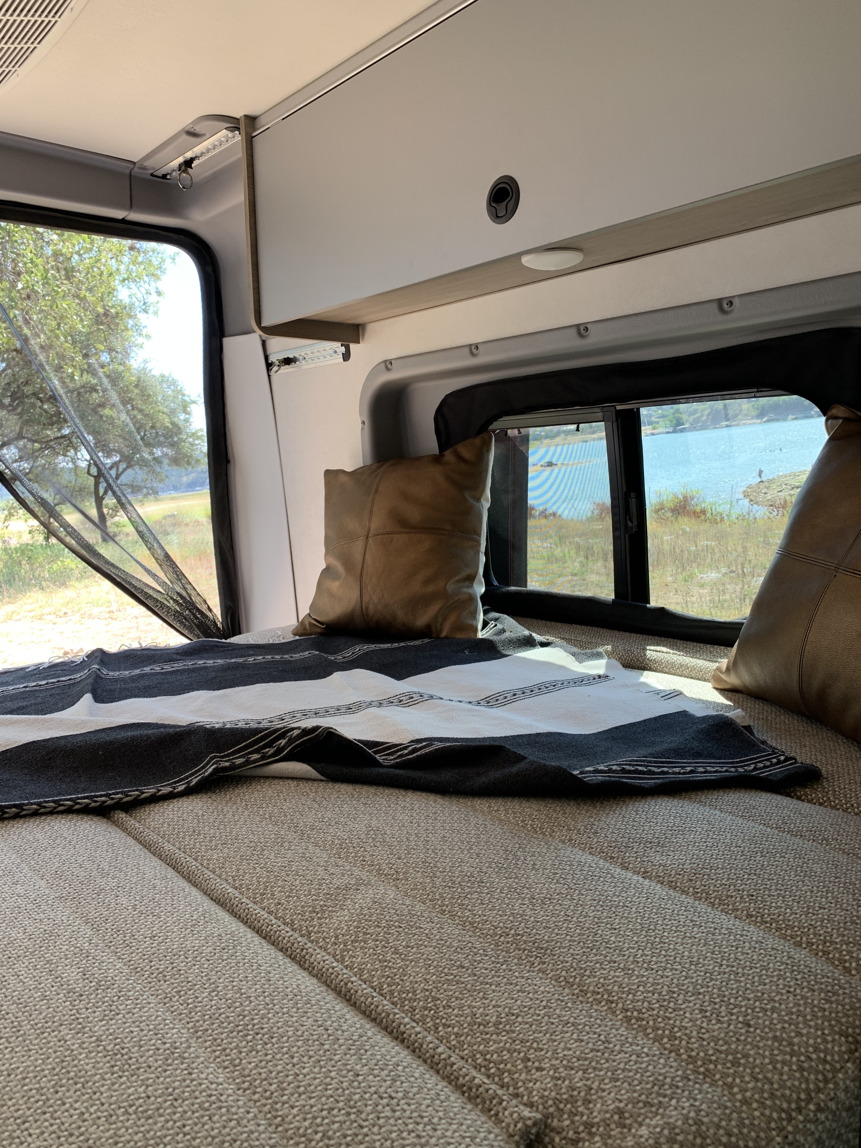 This rv rental offers a comfortable bed with a striped blanket and pillows, overlooking a peaceful lake.. Winnebago Solis 2021