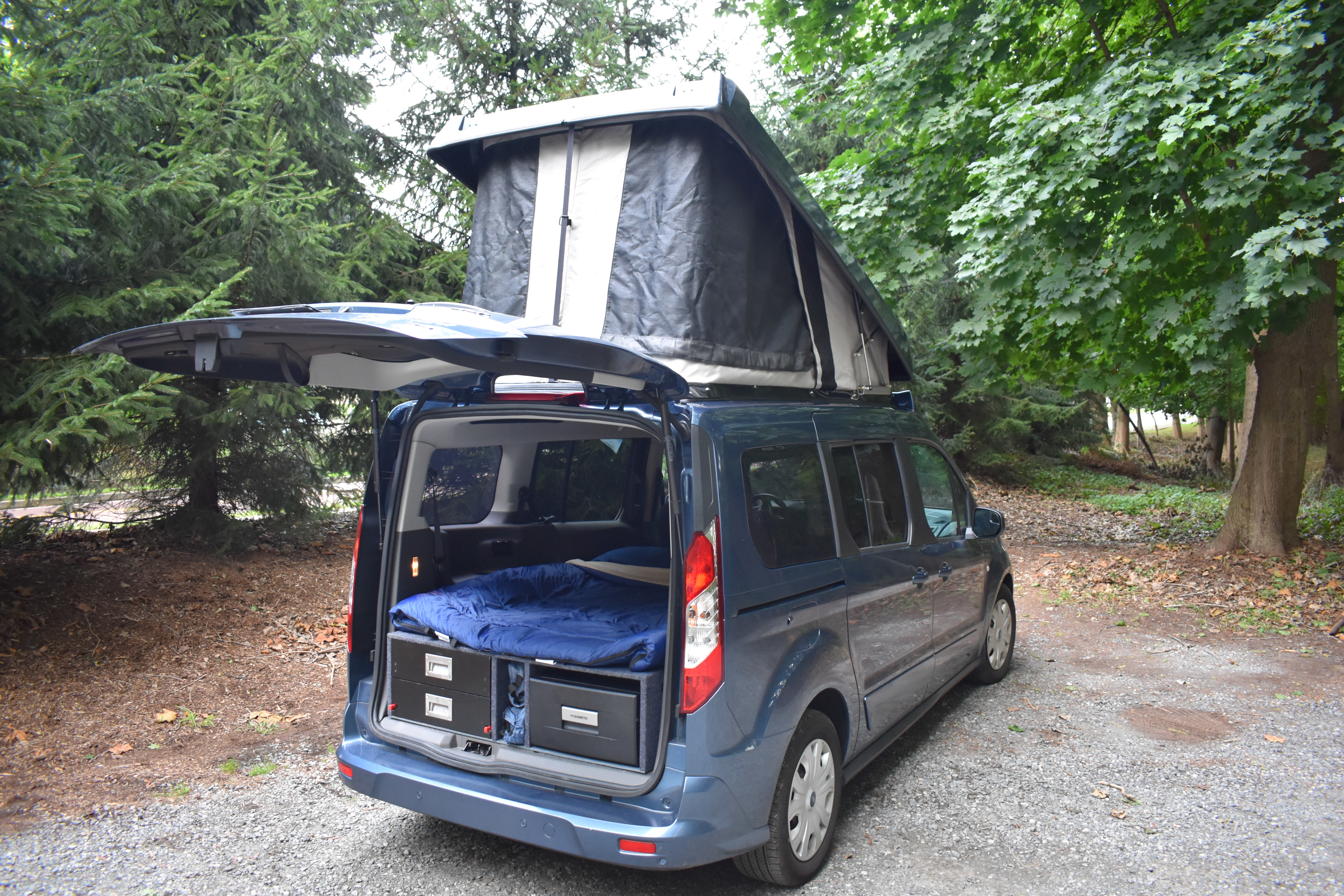 Compact RV rental: A blue pop-top camper van reveals a rear bed, storage drawers, and a Dometic fridge for camping trips.. Ford Van 2019