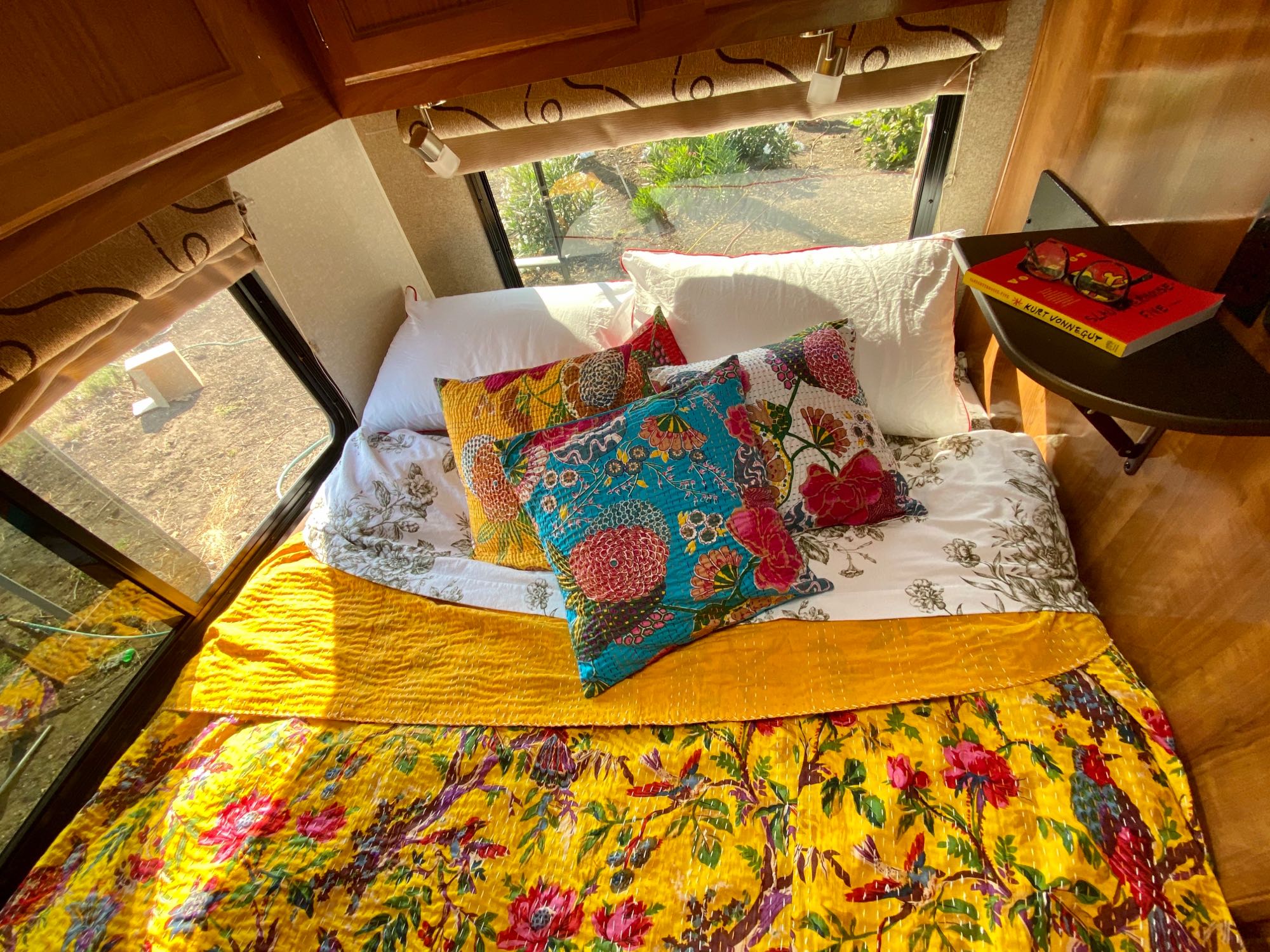 This vibrant RV rental features a cozy bed with yellow, floral bedding, colorful pillows, and a book-ready side table for ultimate comfort on the go.. Coachmen Leprechaun 2019