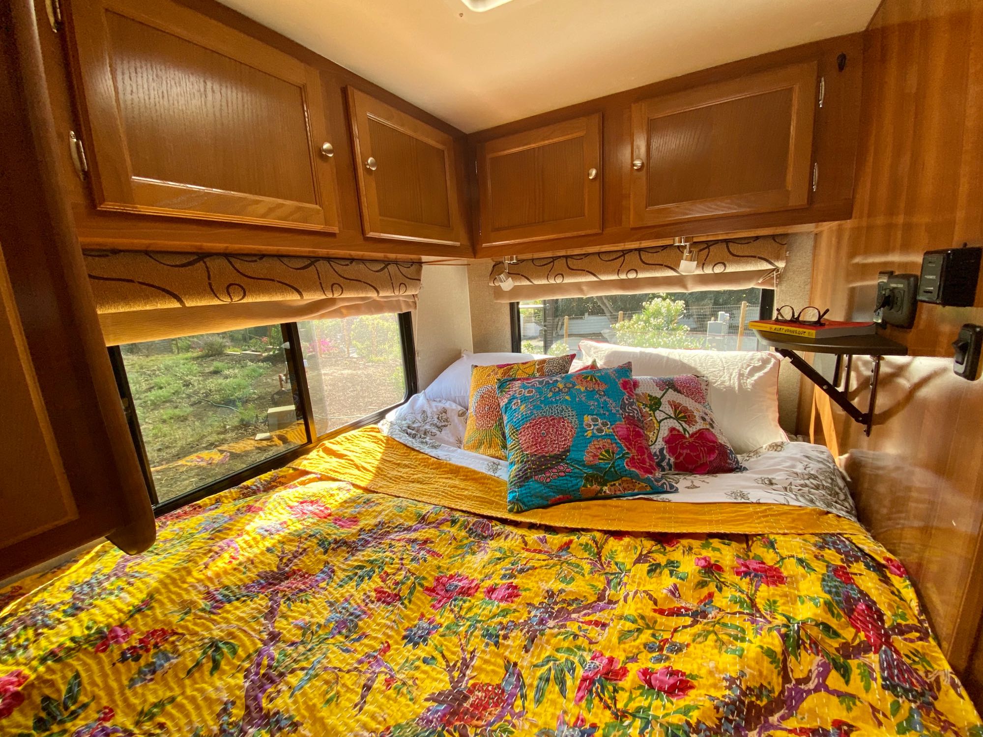 Bright & cozy RV rental bedroom featuring a vibrant yellow floral quilt, colorful pillows, wooden cabinets & large windows.. Coachmen Leprechaun 2019