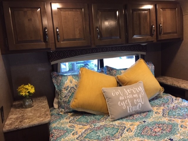 Inviting RV rental bedroom: comfy bed with vibrant pillows, wood cabinets above, and window view, promising relaxation.. Thor Chateau 2017
