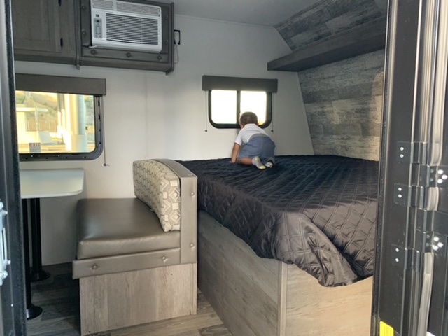 Inside a compact RV rental, a child explores the bed next to a dinette. An AC unit is mounted above.. Keystone Springdale 2021