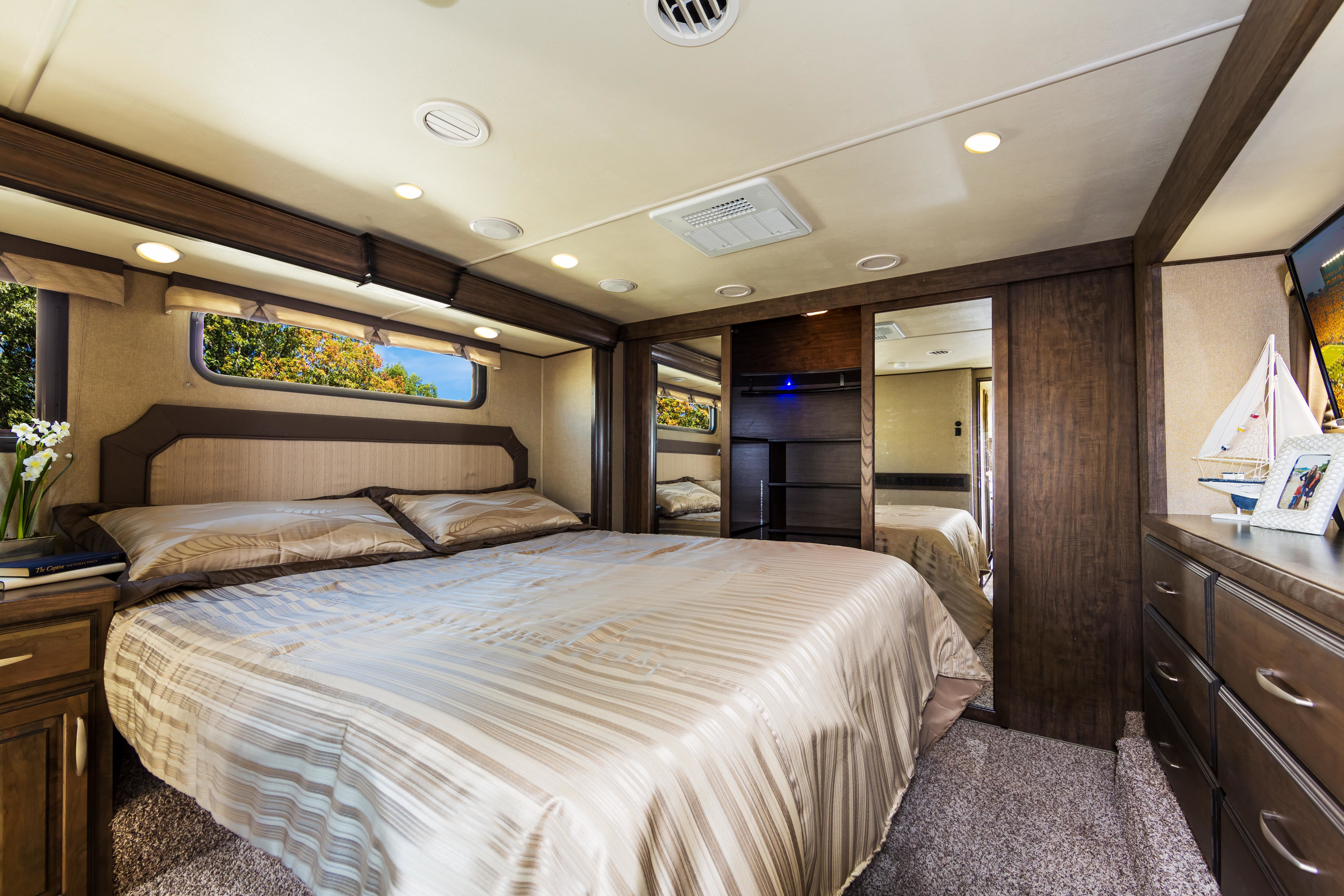 Comfortable RV rental bedroom with a queen bed, dark wood furniture, and windows showing autumn trees. Your adventure awaits.. Grand Design Solitude 2016