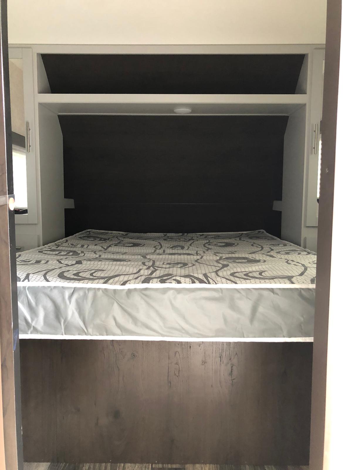View of an RV rental bedroom showing a bed with patterned mattress, dark headboard, and overhead storage.. Keystone Bullet 2020