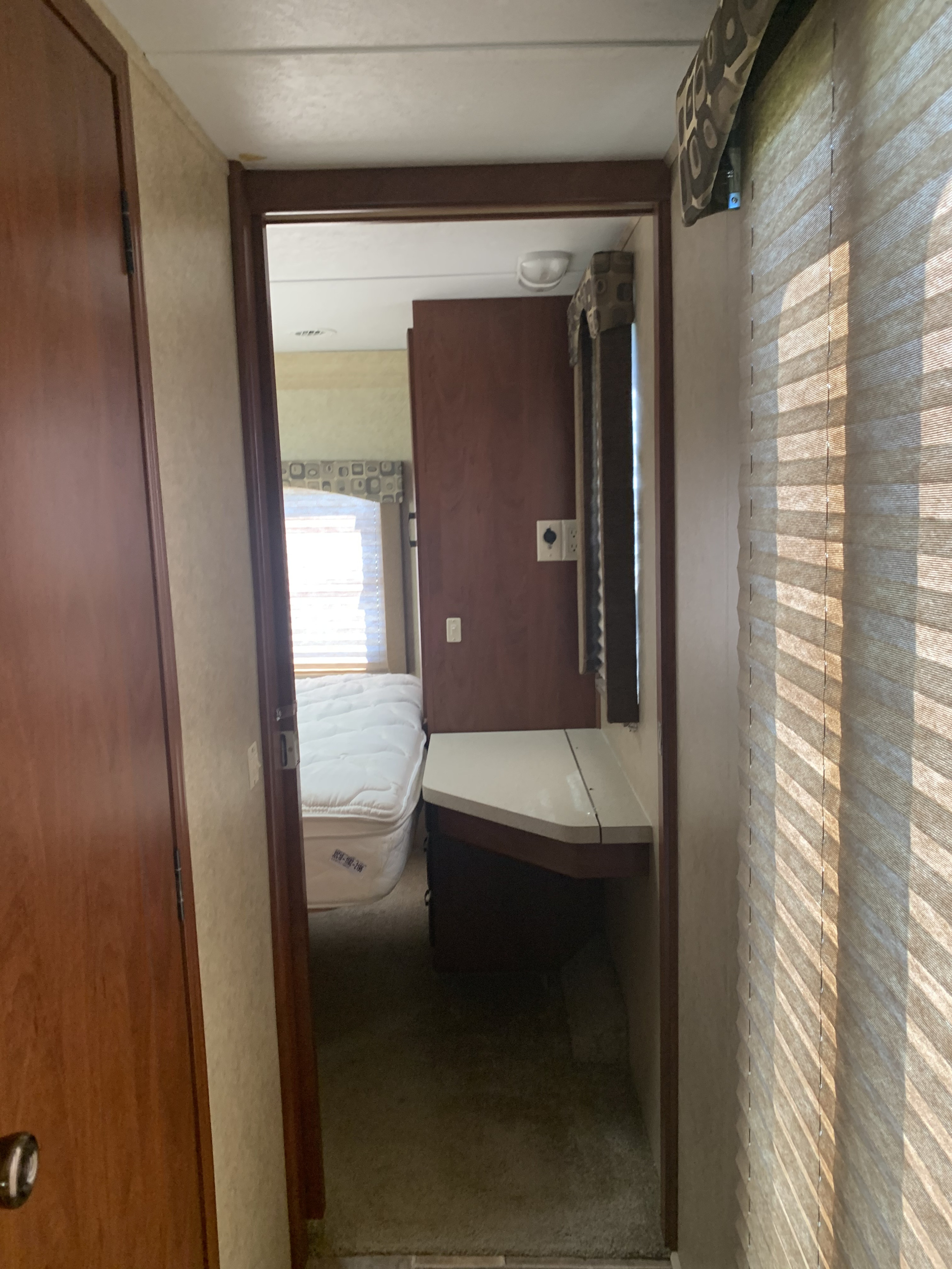 Inside an RV rental, looking into a bedroom with a white mattress, vanity, and window with blinds. A ceiling stain is visible.. Forest River Sunseeker 2012