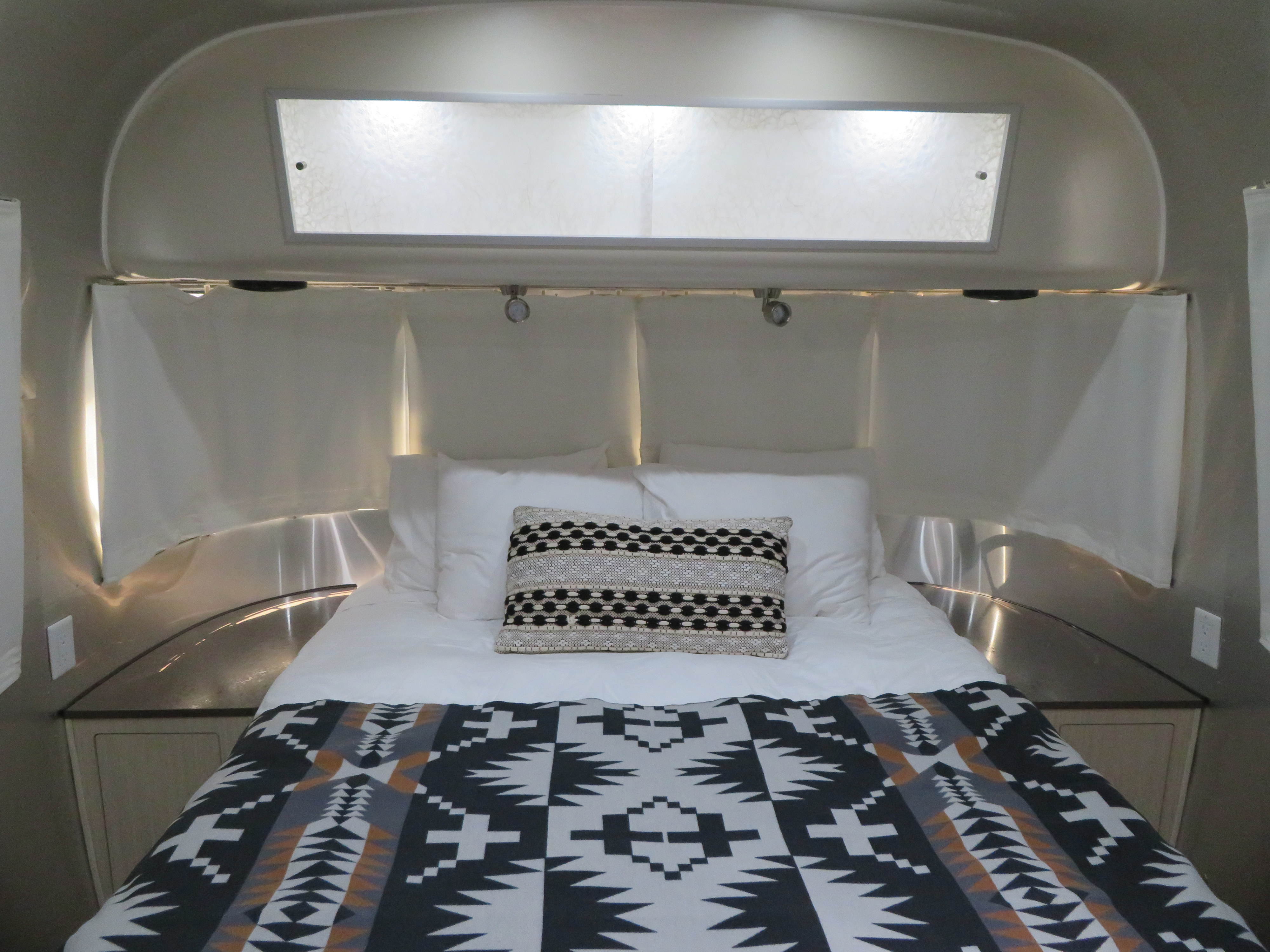 Modern RV rental sleeping area. A cozy bed with white linens and a patterned throw blanket sits within the curved metal interior.. Airstream International 2017