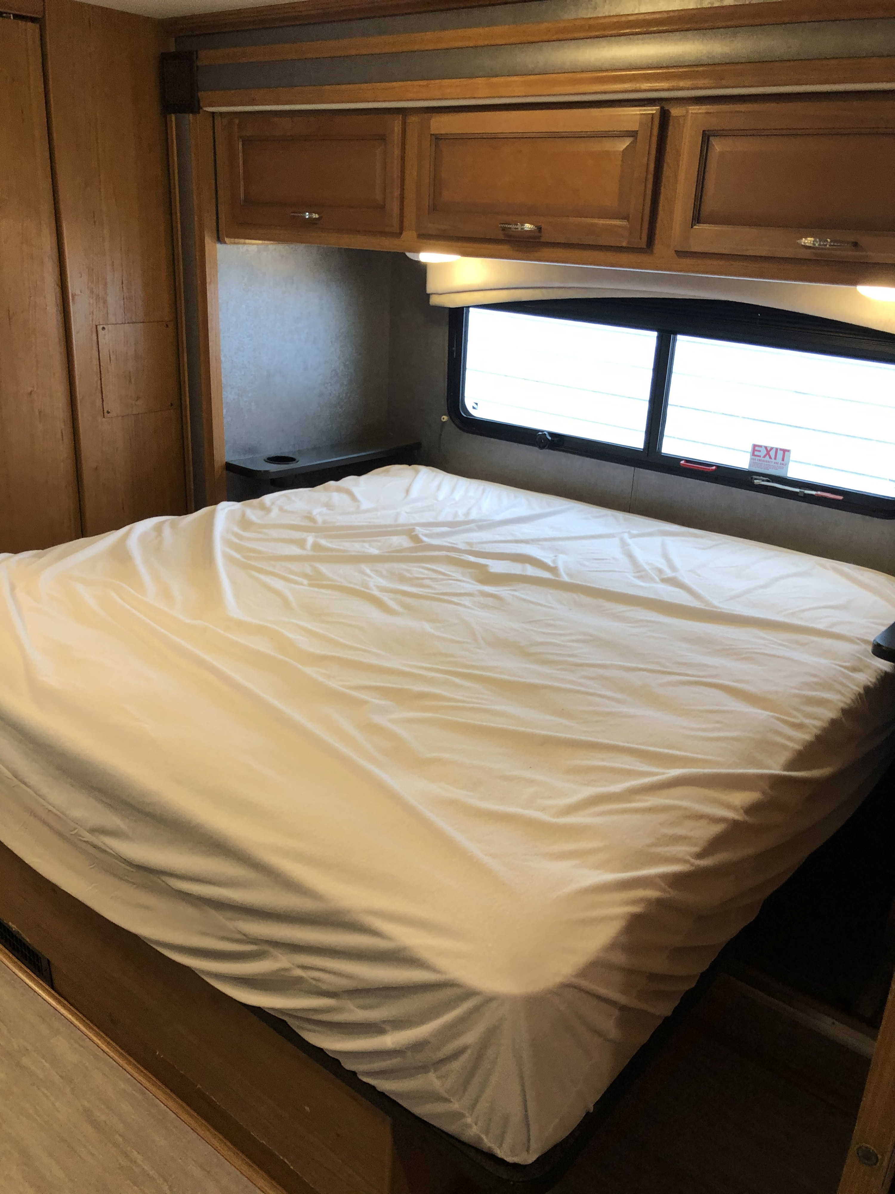 RV rental bedroom showcasing a bed with white sheets, wooden overhead cabinets, and an emergency exit window.. Fleetwood Bounder 2015