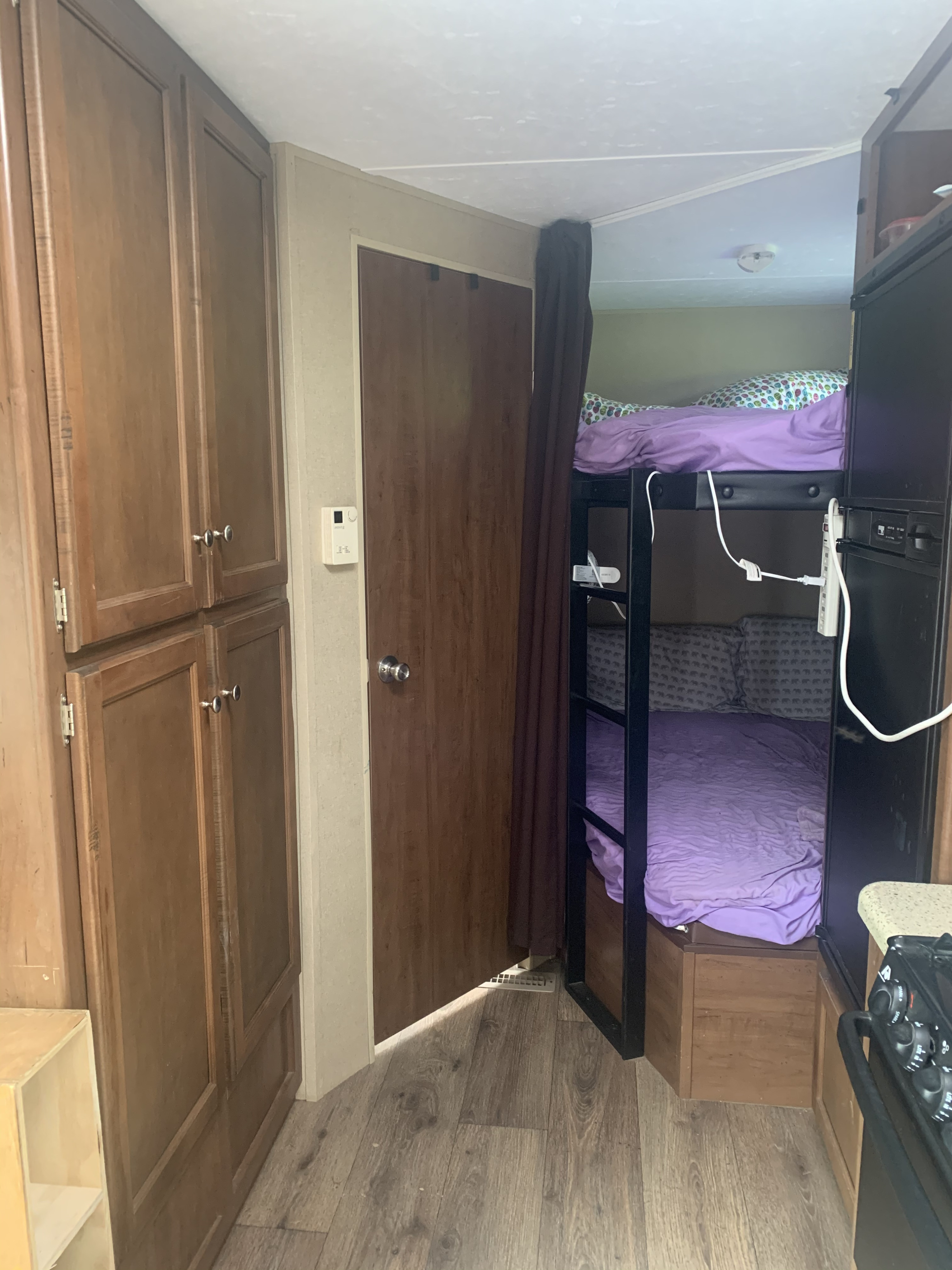 Compact RV rental interior shows wooden storage cabinets, a door, and bunk beds with purple bedding near a small kitchen.. Dutchmen Aspen Trail 2014