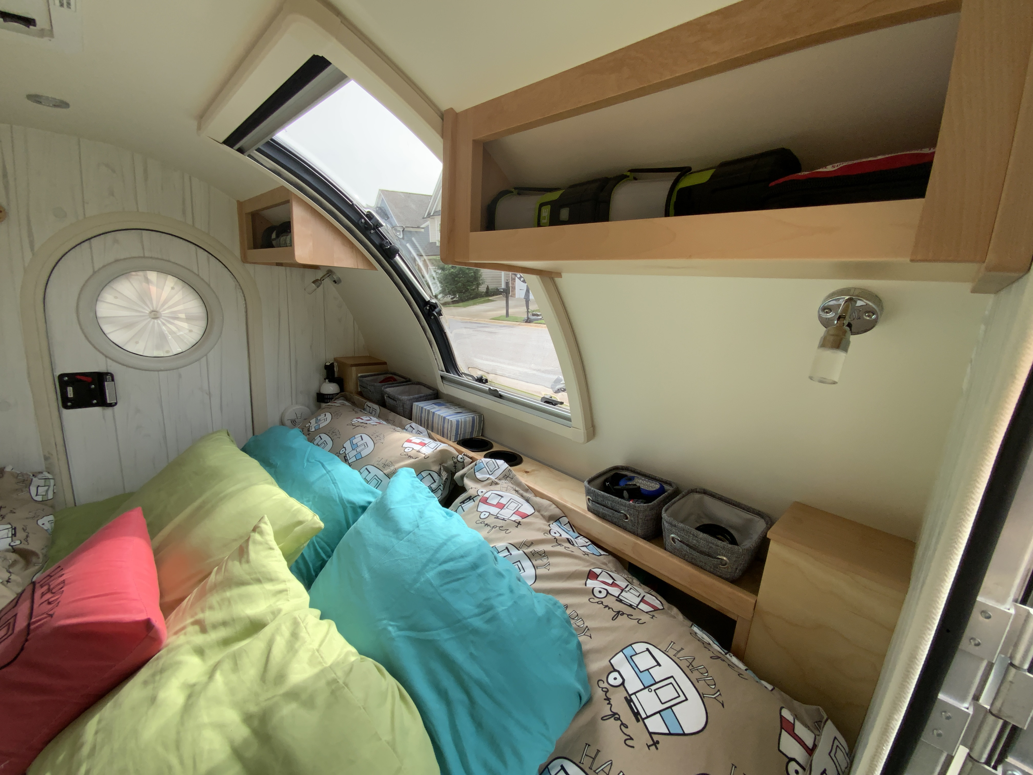 Cozy RV rental bedroom featuring a bed with colorful pillows and "Happy Camper" bedding, bright window, and storage.. nuCamp T@G 2019