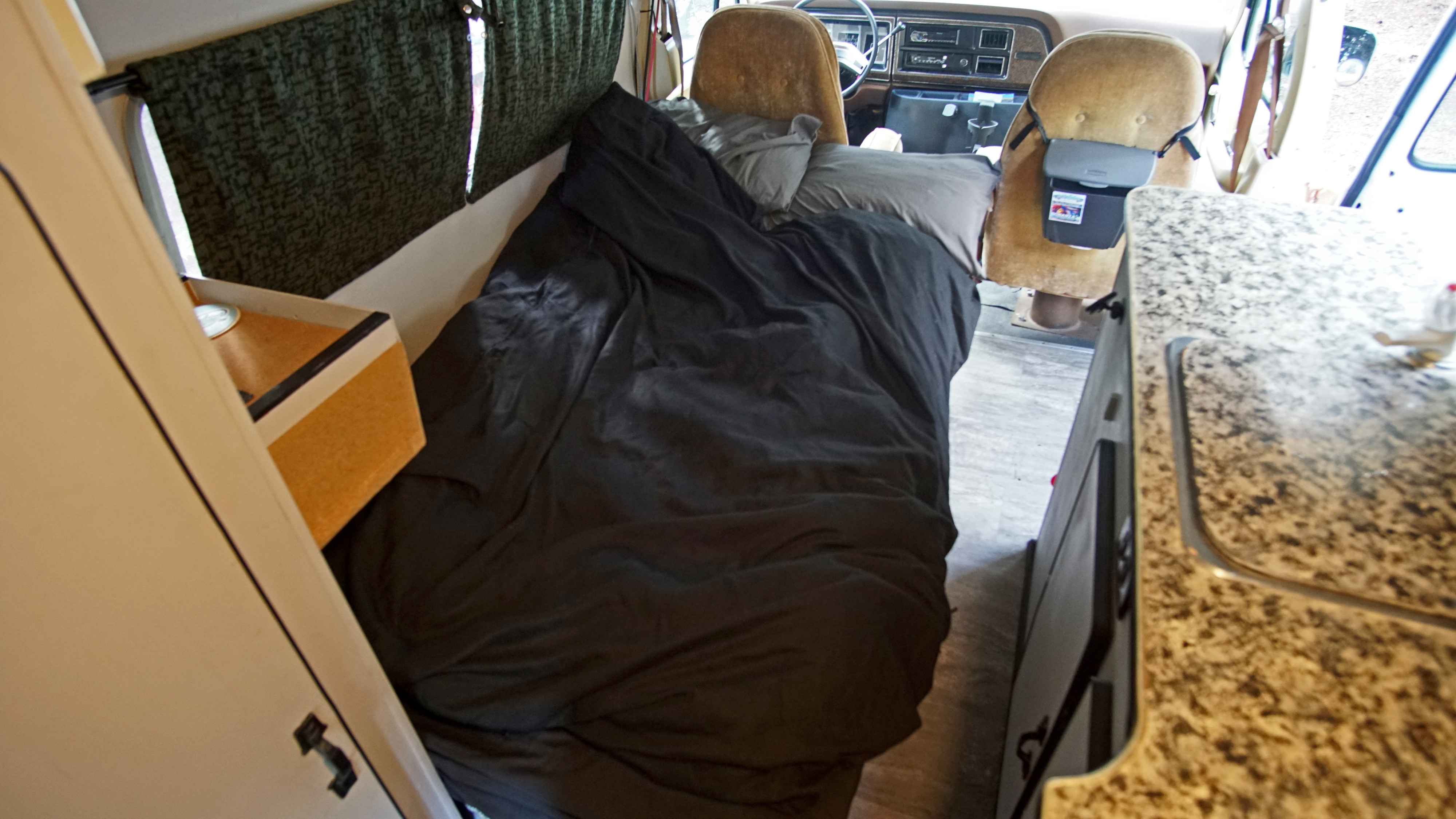 Cozy rv rental interior featuring a bed with black bedding, front cabin seats, and a granite-look kitchen counter.. Ford Econoline Club Wagon 1980