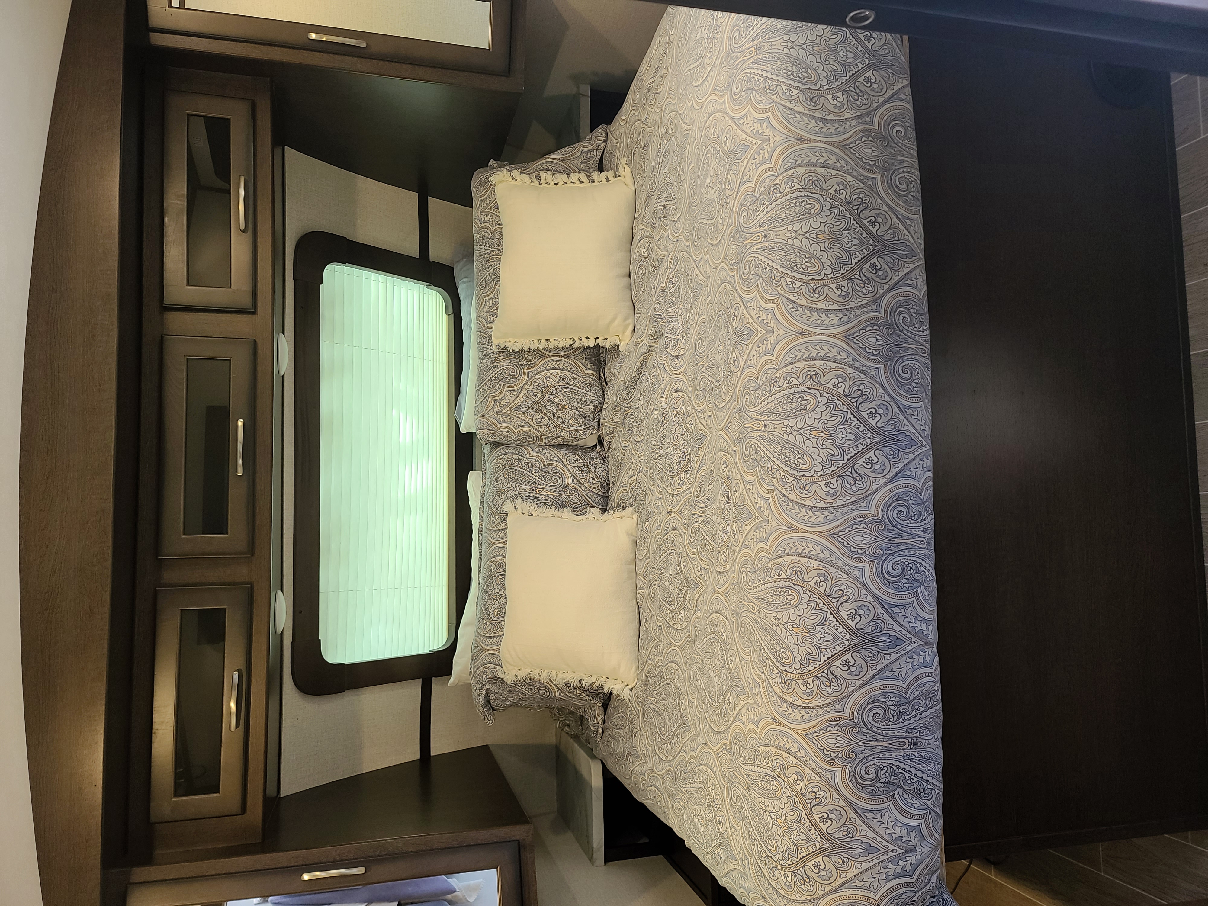Comfortable RV rental bedroom featuring a bed with patterned paisley bedding, white pillows, and built-in wood cabinets and window.. Dutchmen Other 2020
