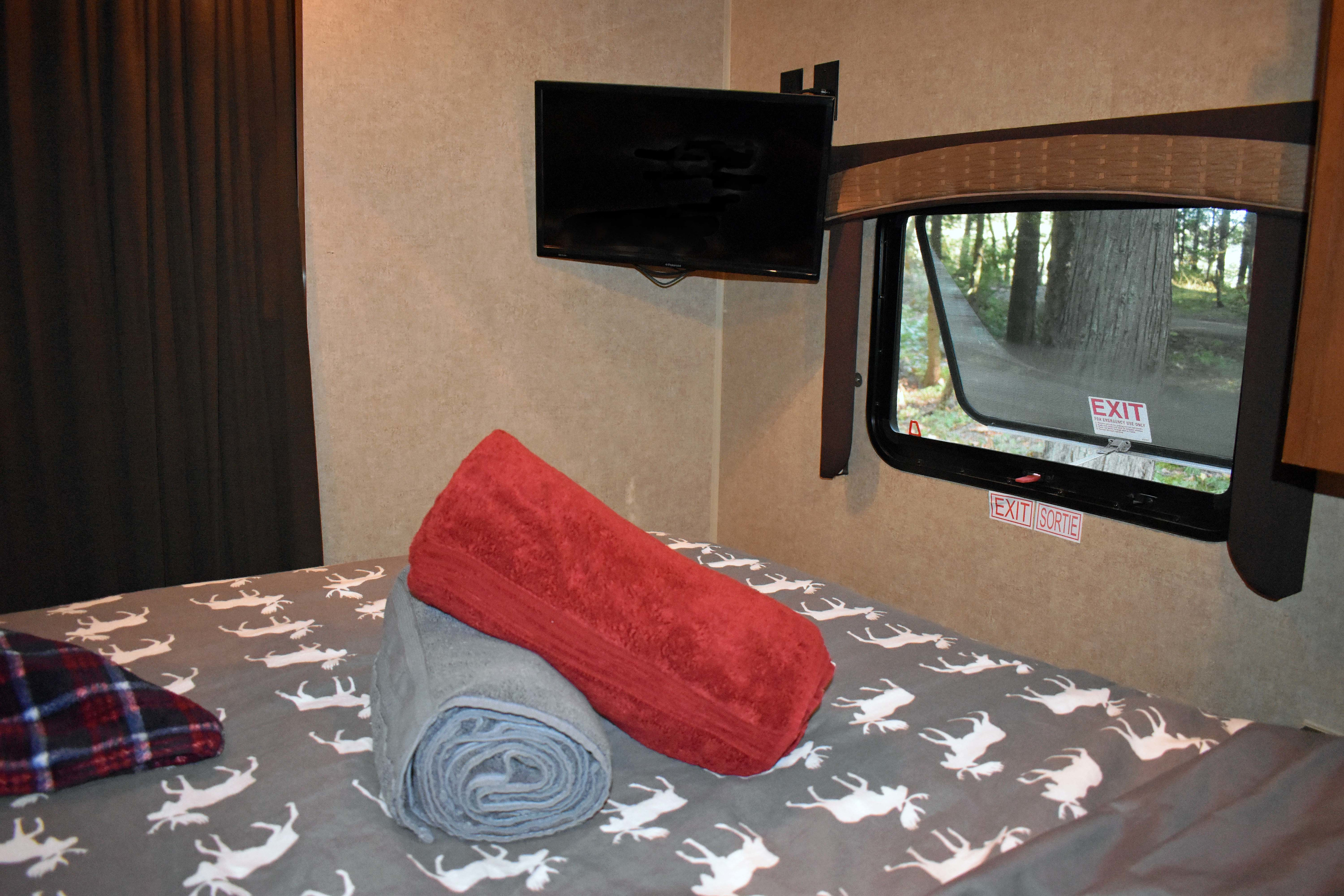 Cozy RV rental bedroom with deer-patterned bed, towels, TV, and an emergency exit window overlooking trees.. Jayco Jay Flight 2014