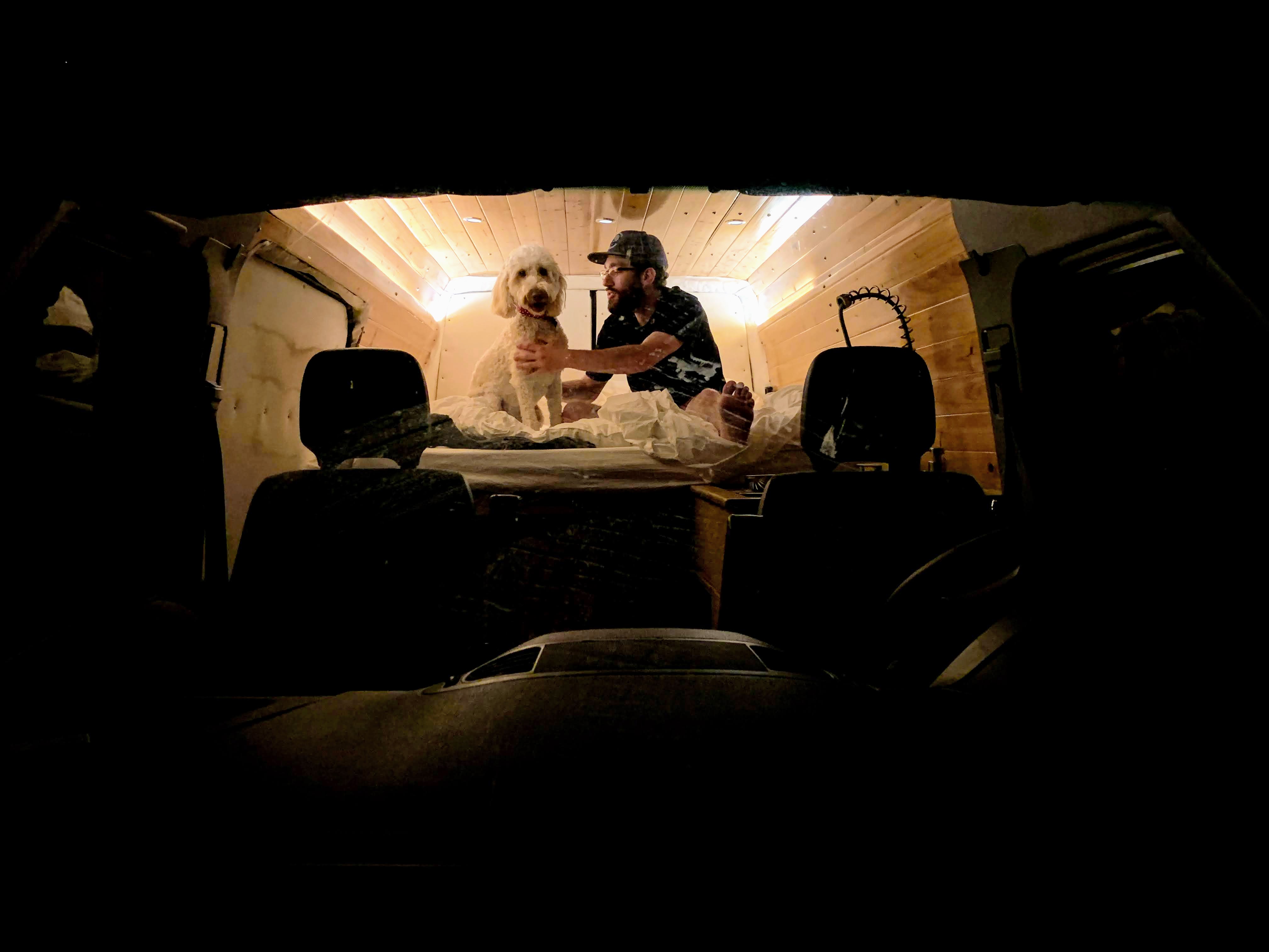 Cozy RV rental interior shows a man and his fluffy dog on a bed with wood paneling, viewed from the driver's seat.. Mercedes Sprinter 2006