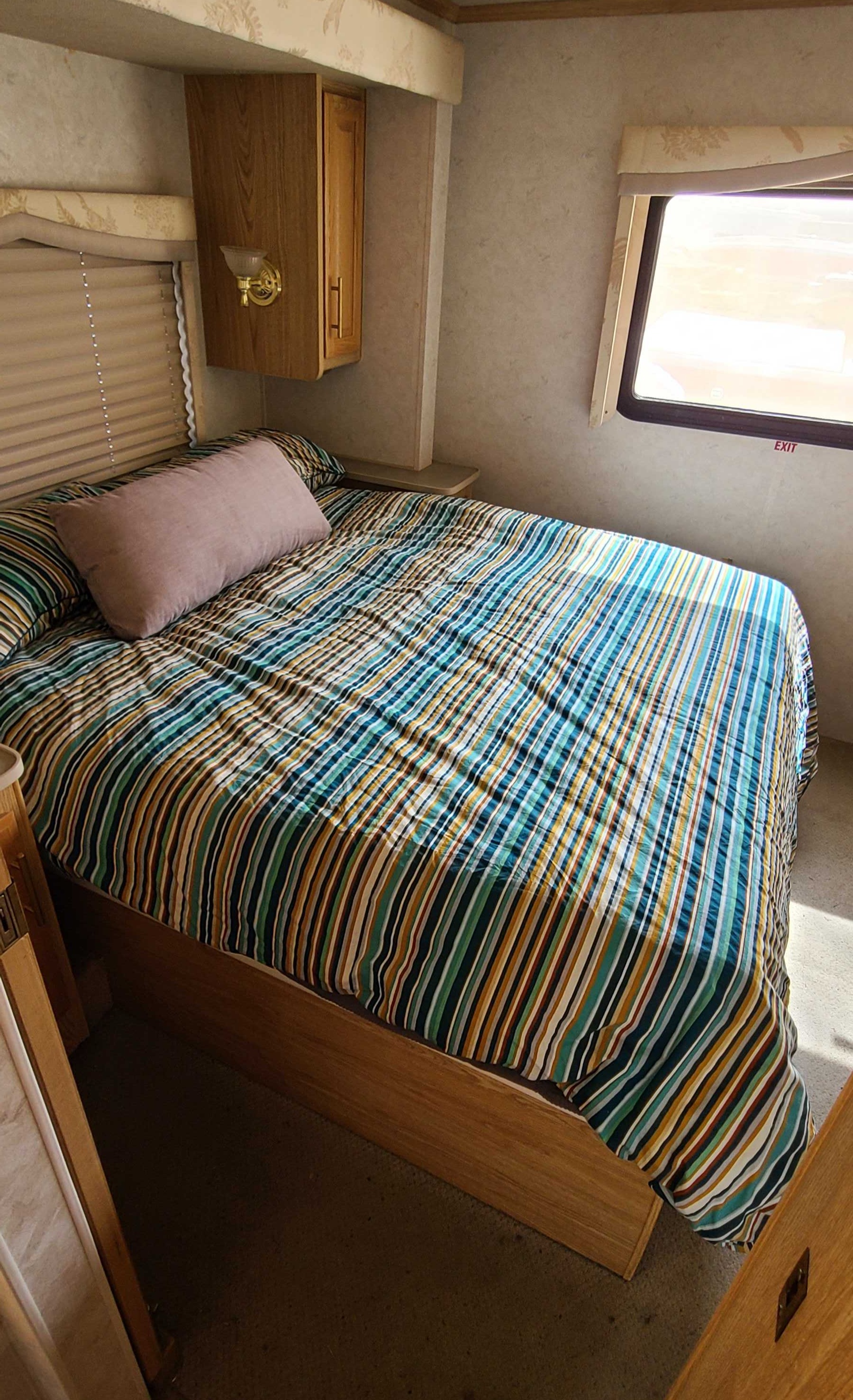 Cozy RV rental bedroom featuring a colorful striped bedspread, small window with 'EXIT' sign, and wood cabinets. Perfect for travel.. Fleetwood Bounder 2002