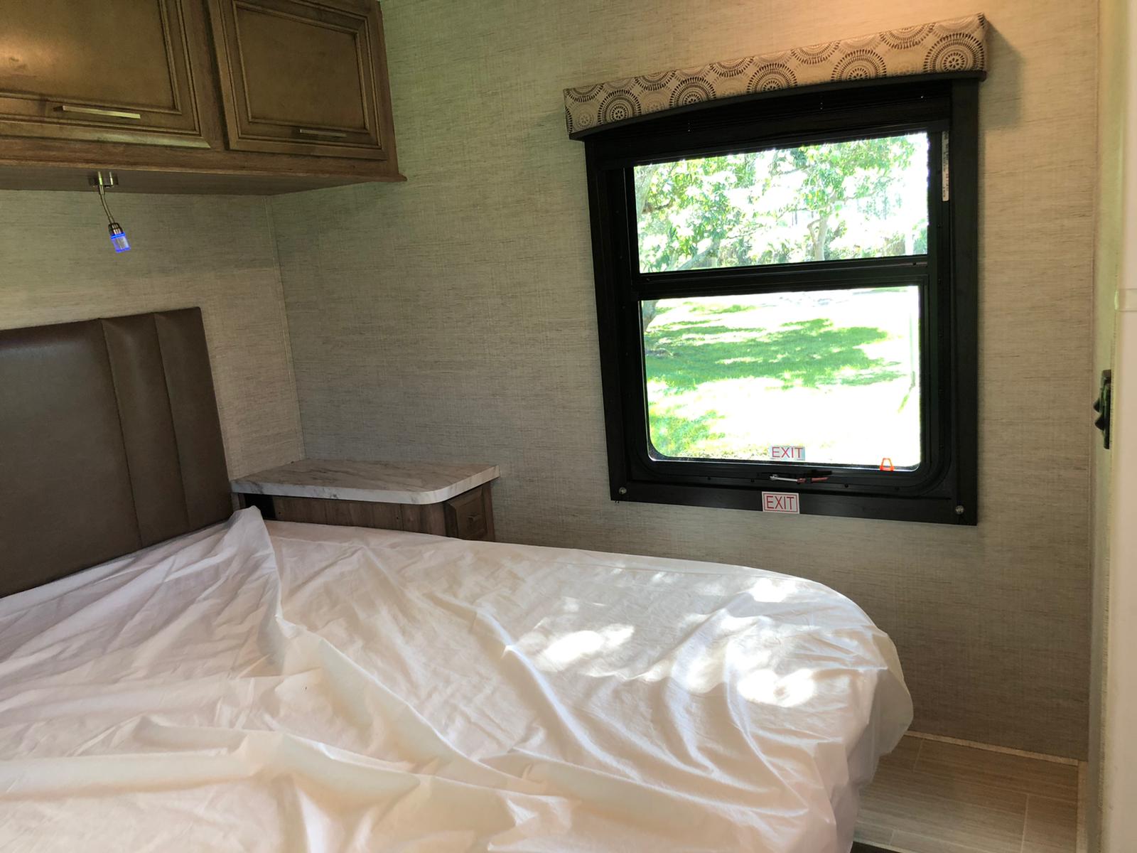 RV rental bedroom: a comfortable bed with white sheets, overhead cabinet, nightstand, and an emergency exit window with green outdoor views.. Entegra Coach Odyssey 29K 2021
