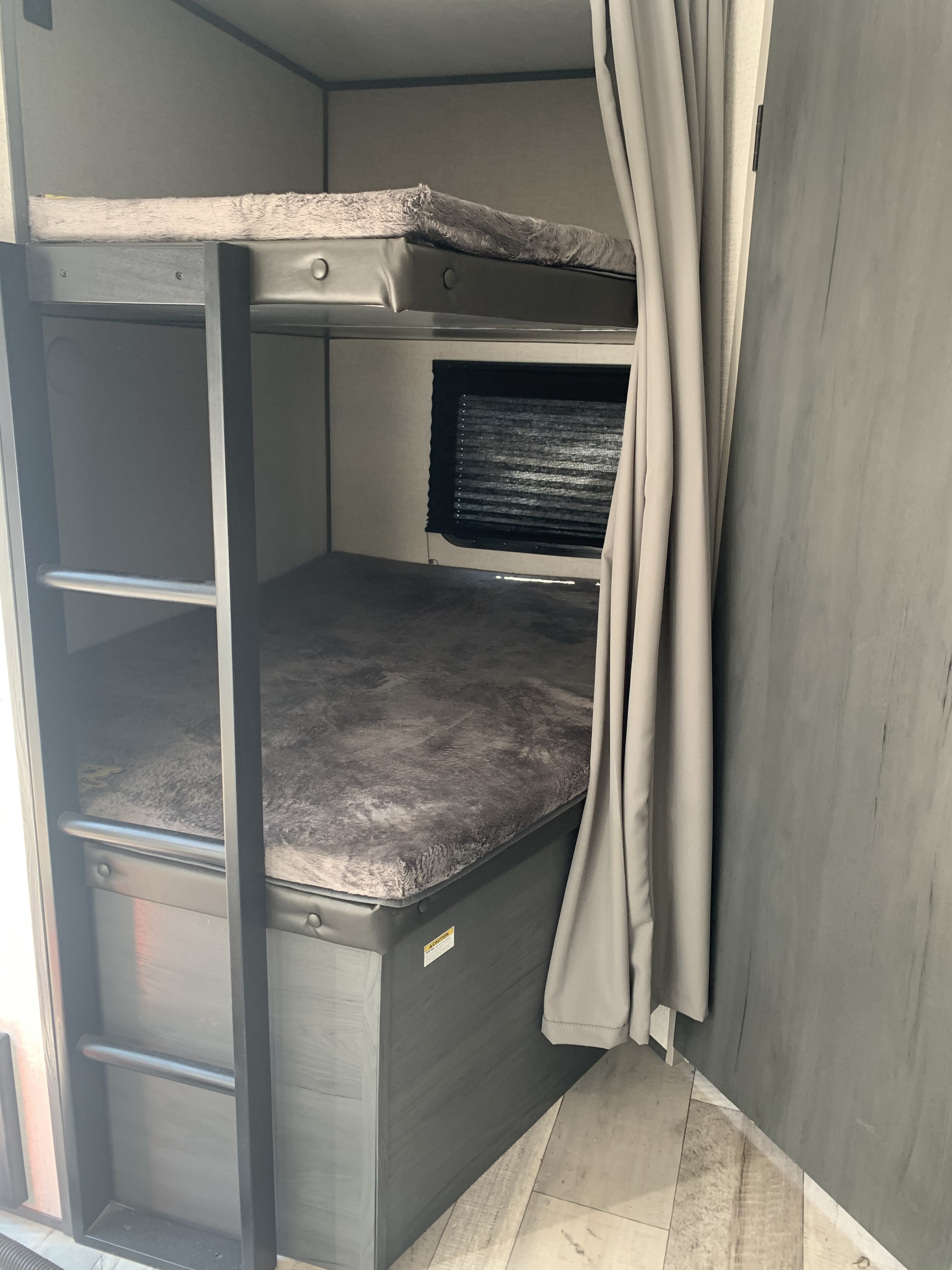 A cozy RV rental interior features stacked bunk beds with soft gray bedding, a window with blinds, and privacy curtains, ideal for family travel.. Dutchmen Aspen Trail 2021