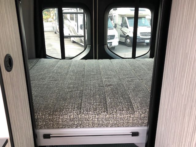 RV rental: Inside a camper van, a comfortable full bed faces rear windows with views of other RVs, ready for travel.. Winnebago Revel 2021