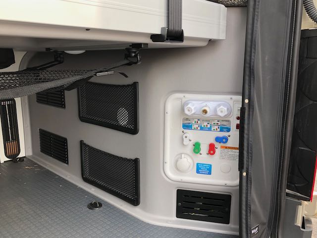 Essential water system panel with hookups and outdoor shower in an RV rental's cargo area, with mesh storage.. Winnebago Revel 2021