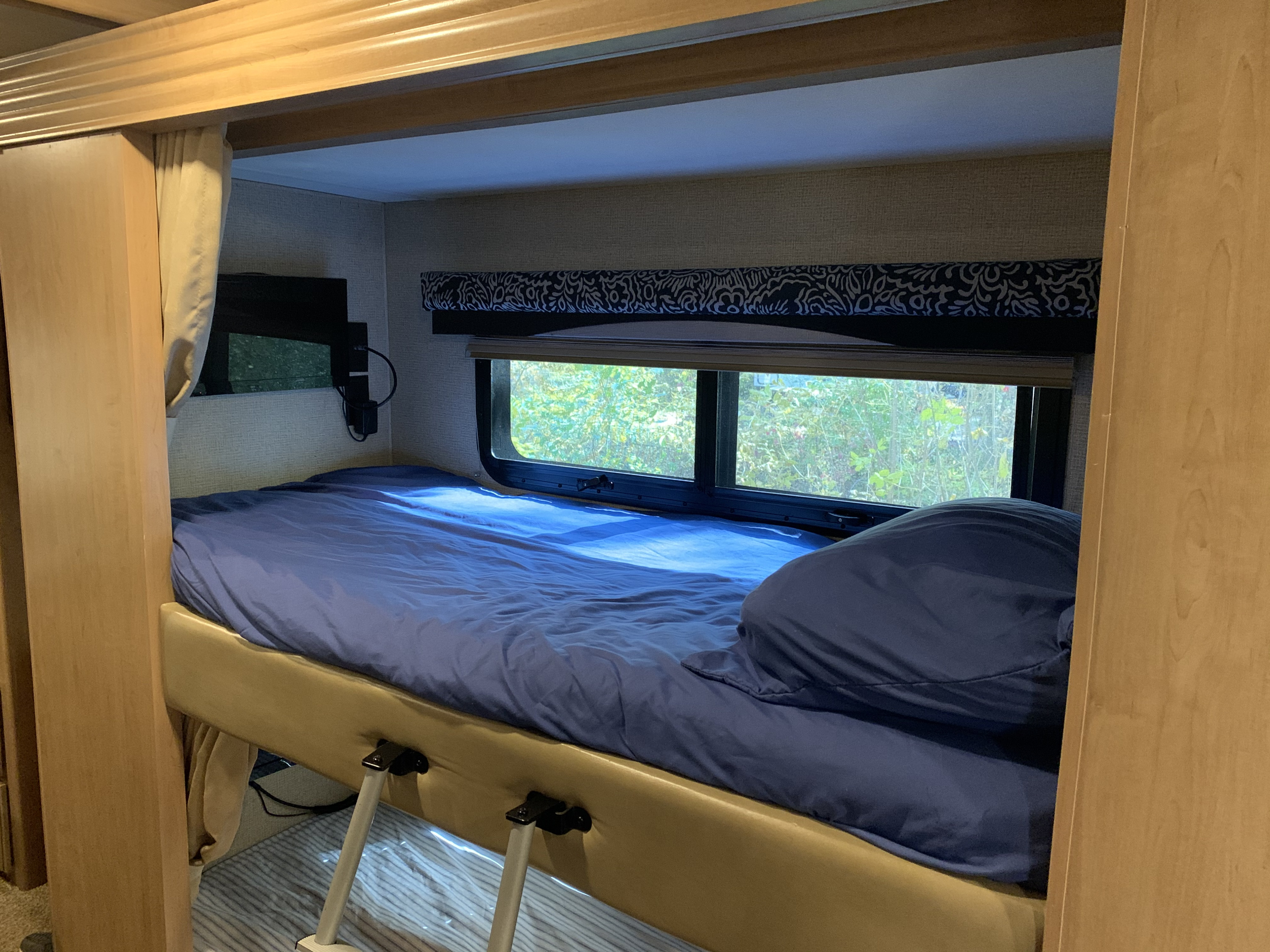 Cozy RV rental bunk bed setup with blue bedding, a window overlooking trees, and a small TV. Ideal for travels.. Thor A.C.E. 2018