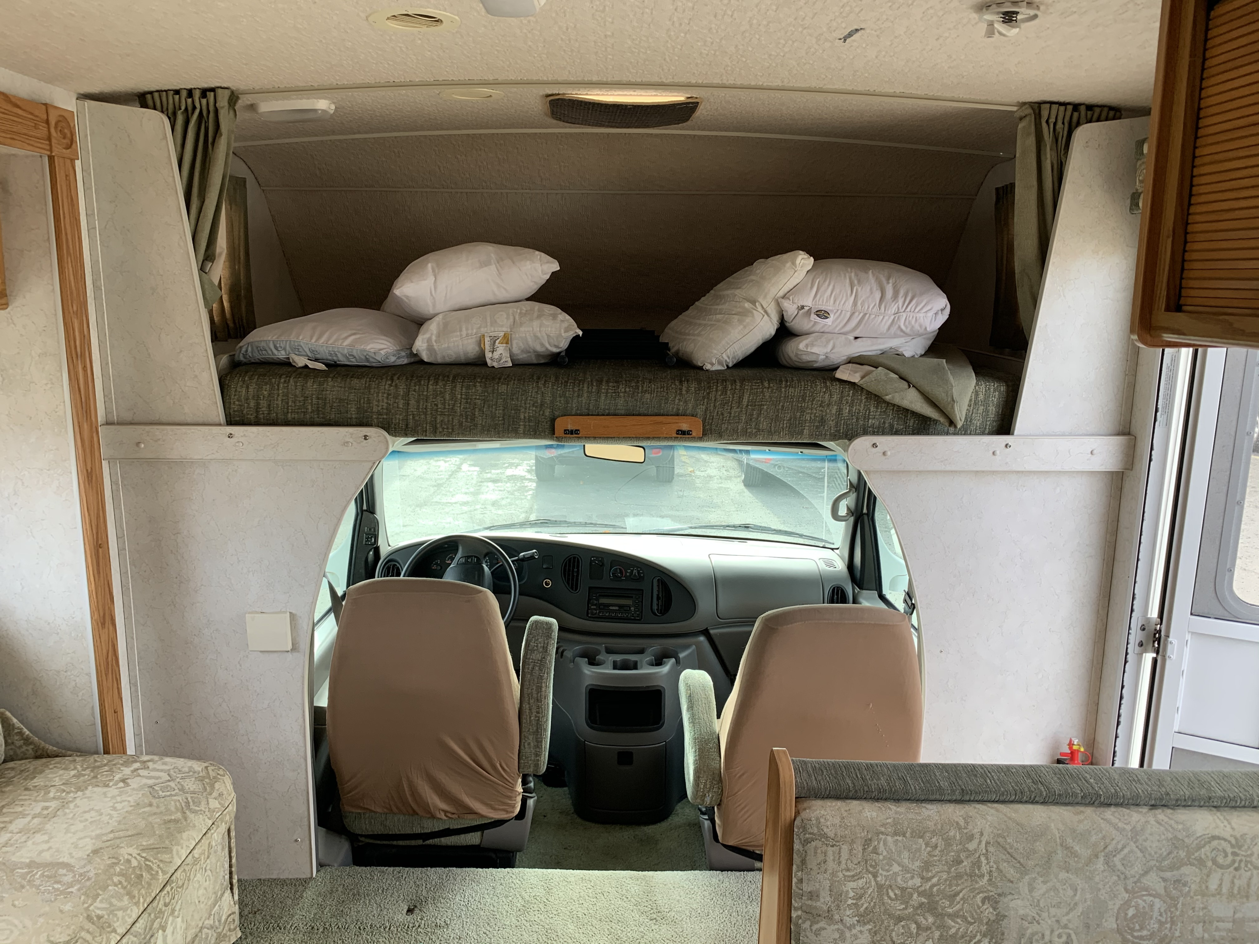 Spacious RV rental interior: front cab with driver's seat, dashboard, and an overhead bunk with pillows.. Winnebago Minnie Winnie 2003
