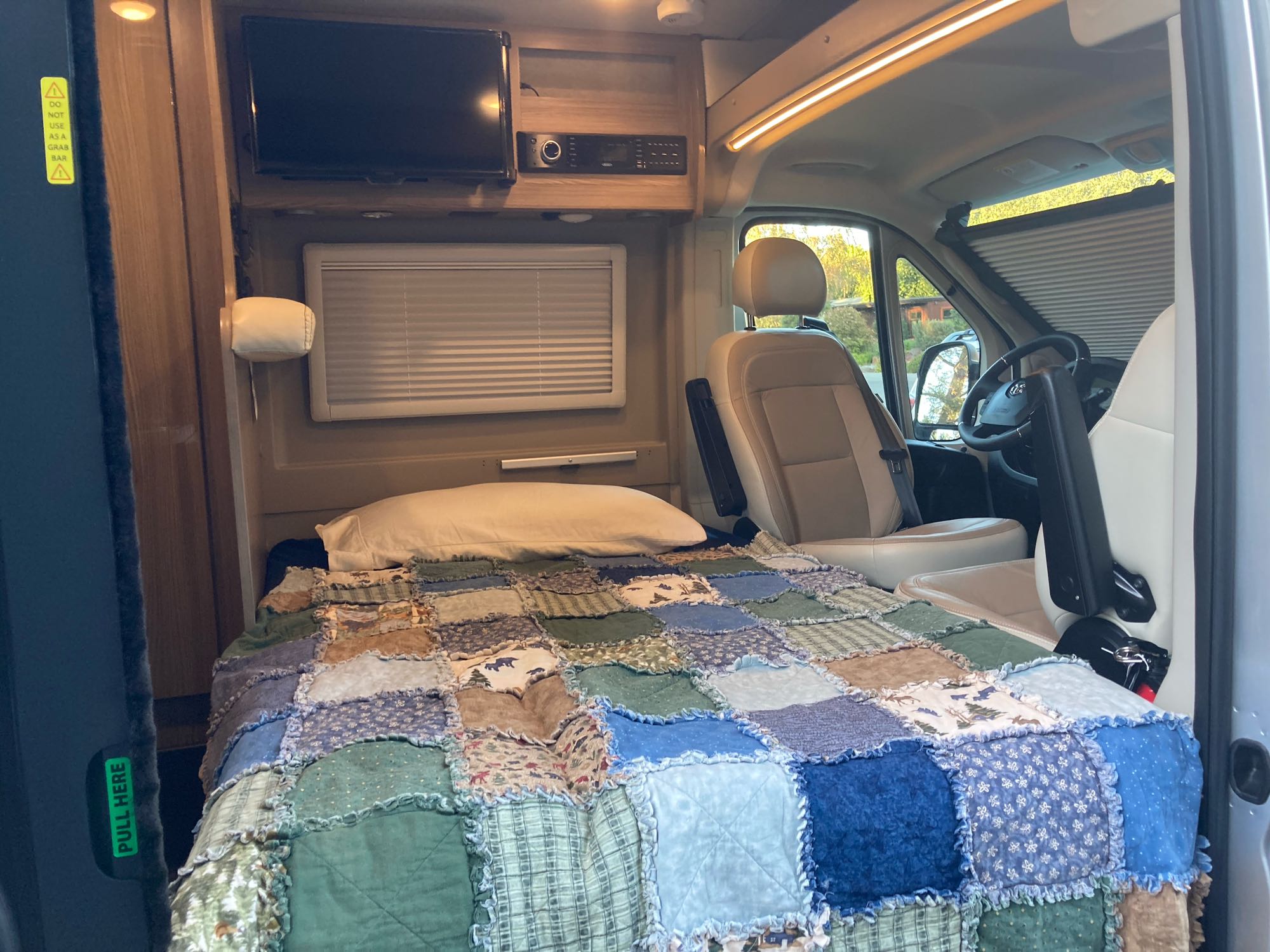 RV rental interior: cozy bed with patchwork quilt, TV, and front cockpit with sunshade.. Winnebago Travato 2020