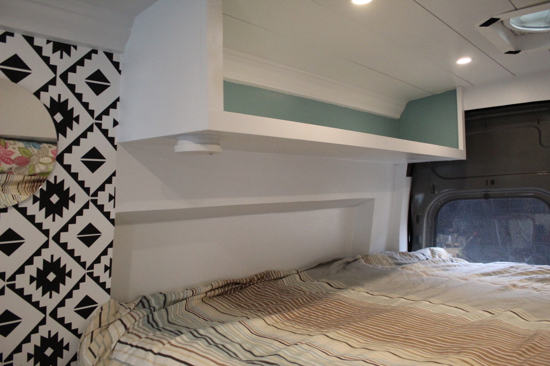 Interior of an RV rental featuring a cozy bed, geometric wall pattern, and integrated overhead storage with accent lighting.. Ford Transit 2020