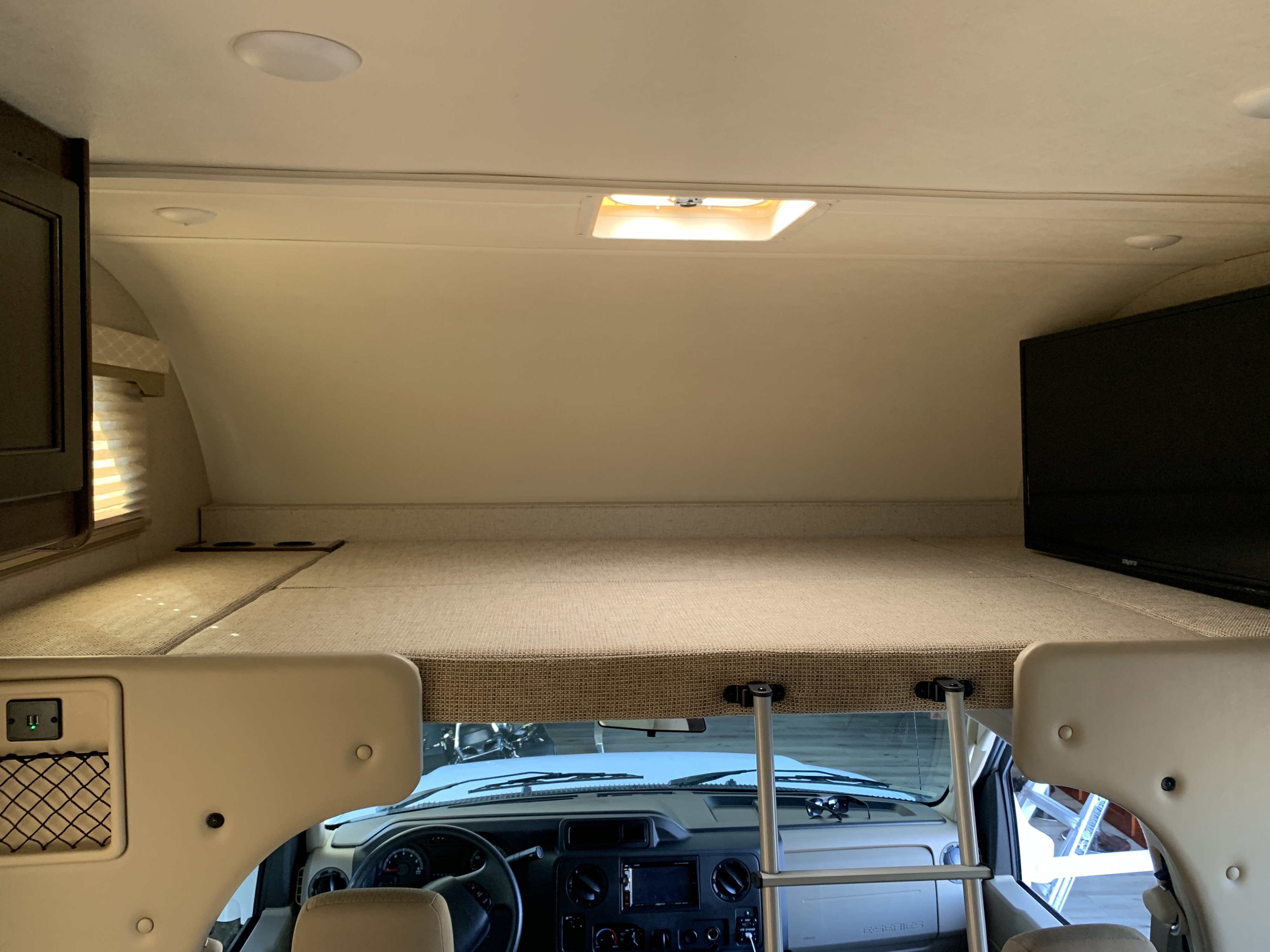 Interior of an rv rental showing the cab-over bunk with bed, TV, and window, overlooking the driver's area.. Thor Freedom Elite 2021