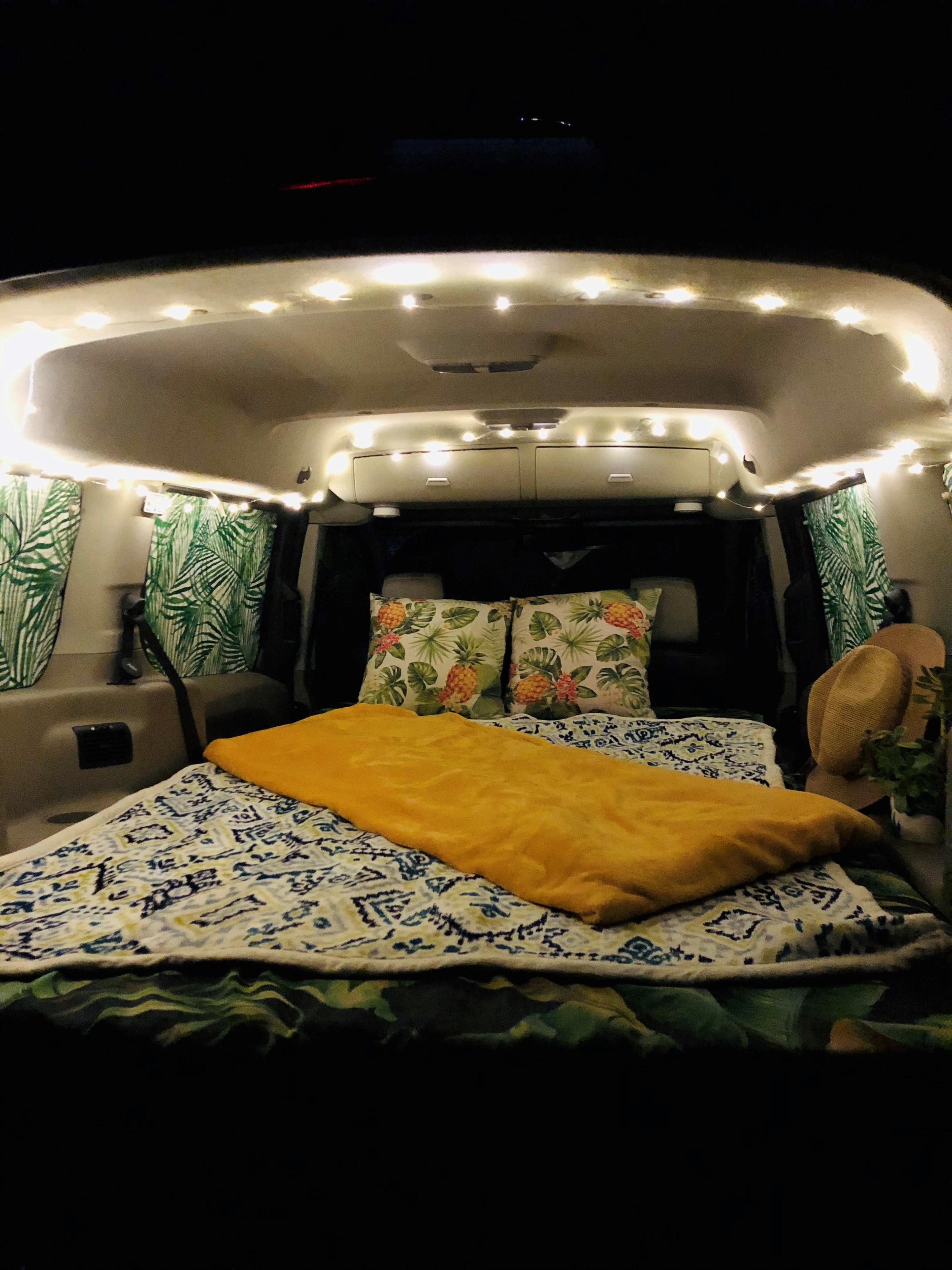 Cozy RV rental interior at night. A comfy bed with patterned blankets, pineapple pillows, and palm-leaf curtains are illuminated by string lights.. Ford transit 2015