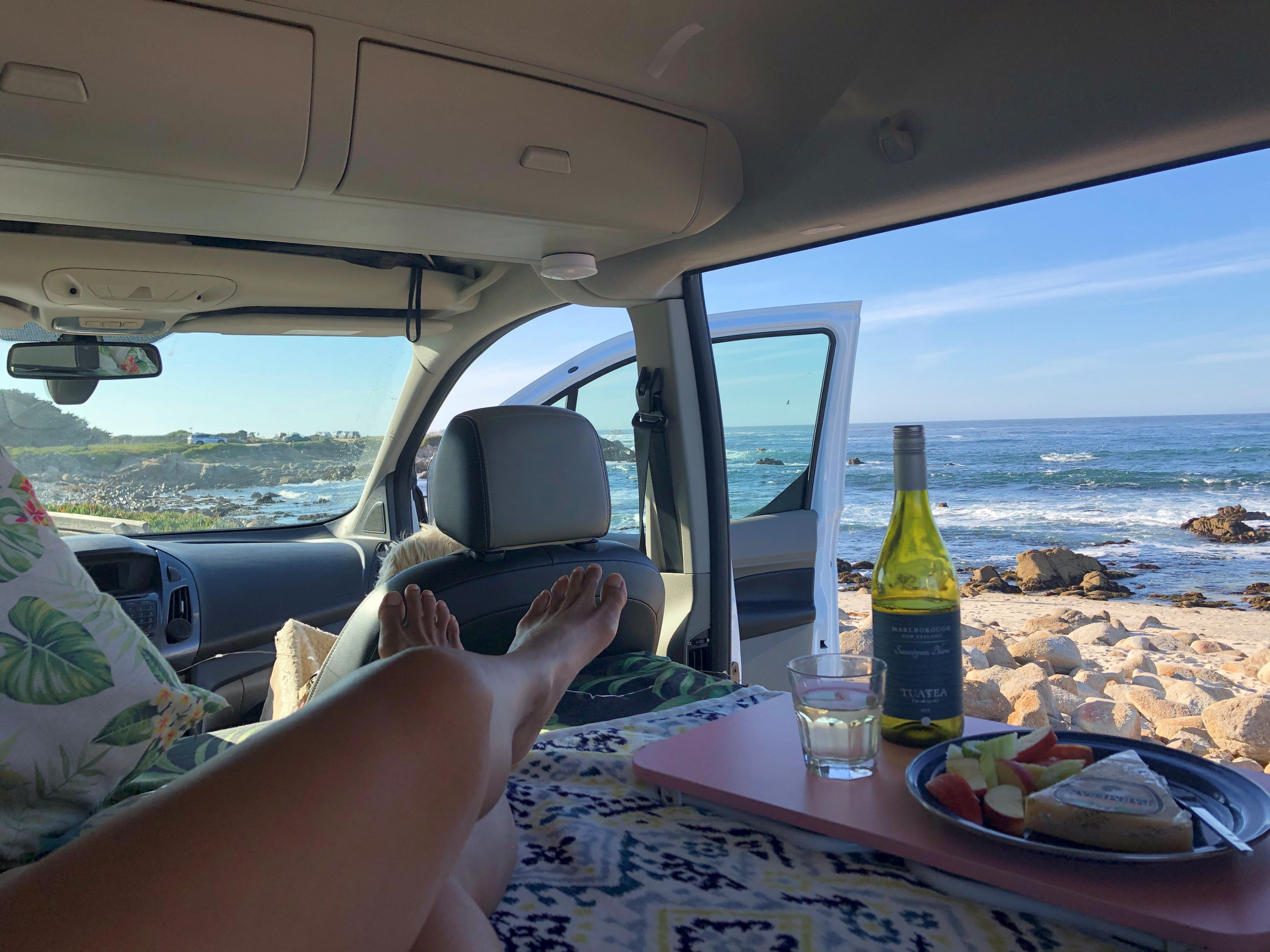 A person relaxes in an RV rental, feet up, enjoying wine, snacks, and ocean views from inside their campervan.. Ford transit 2015
