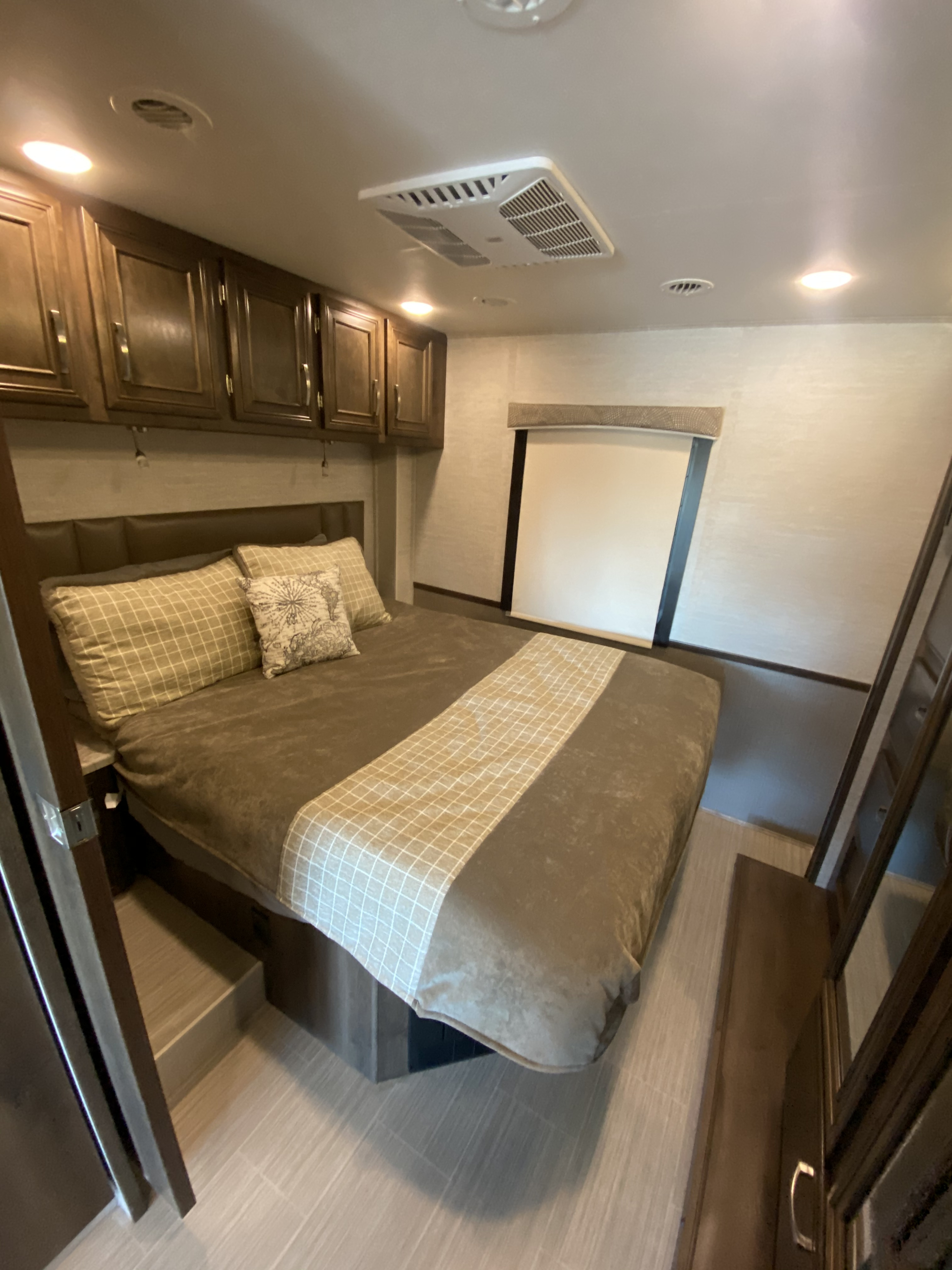 Comfortable RV rental bedroom featuring a large bed, patterned bedding, overhead wooden cabinets, and a ceiling AC unit.. Entegra Coach Other 2021