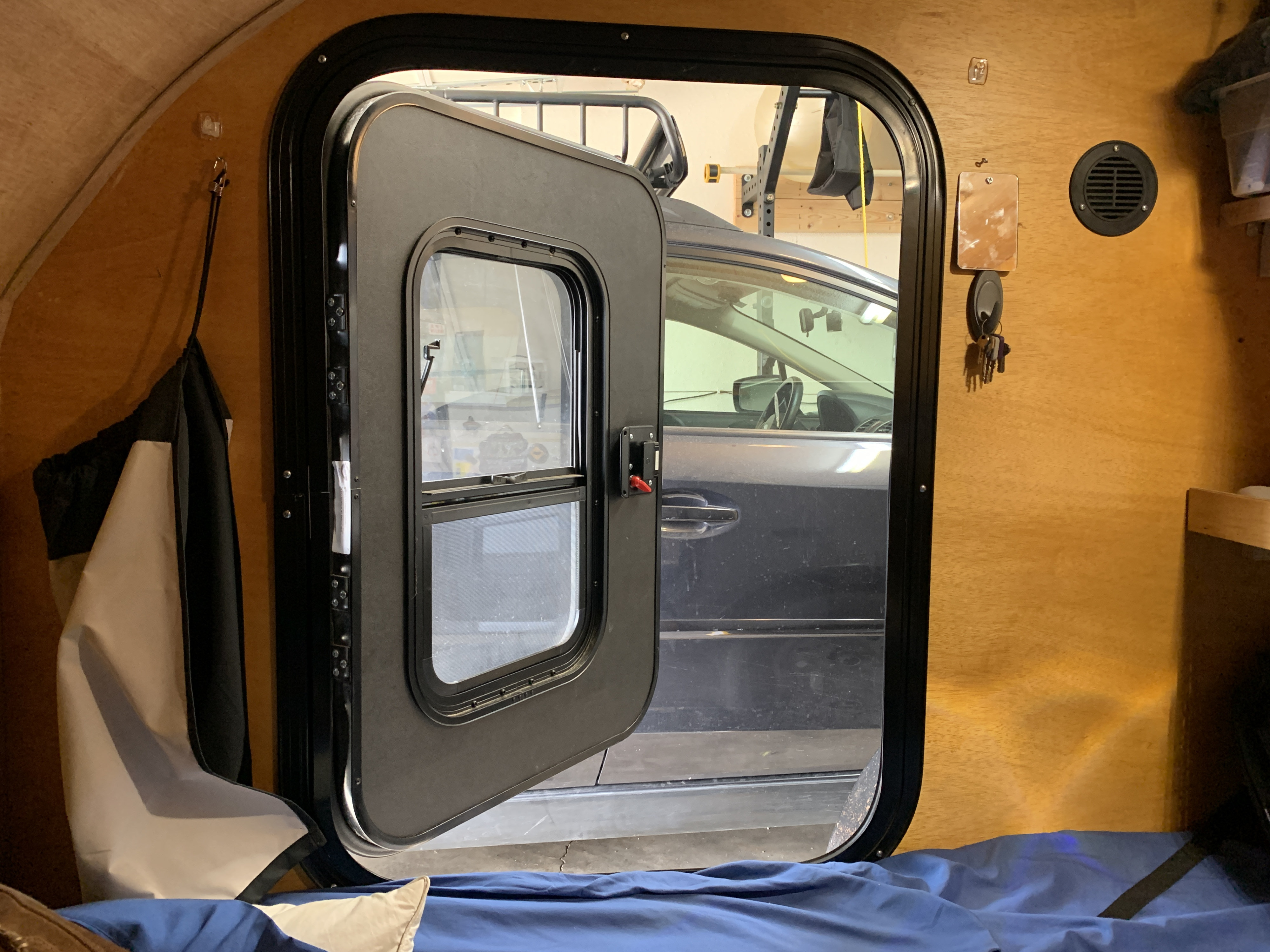 Inside a cozy rv rental, this camper's plywood interior features a bed, hooks, and an open door to a parked car.. Little Guy Other 2020