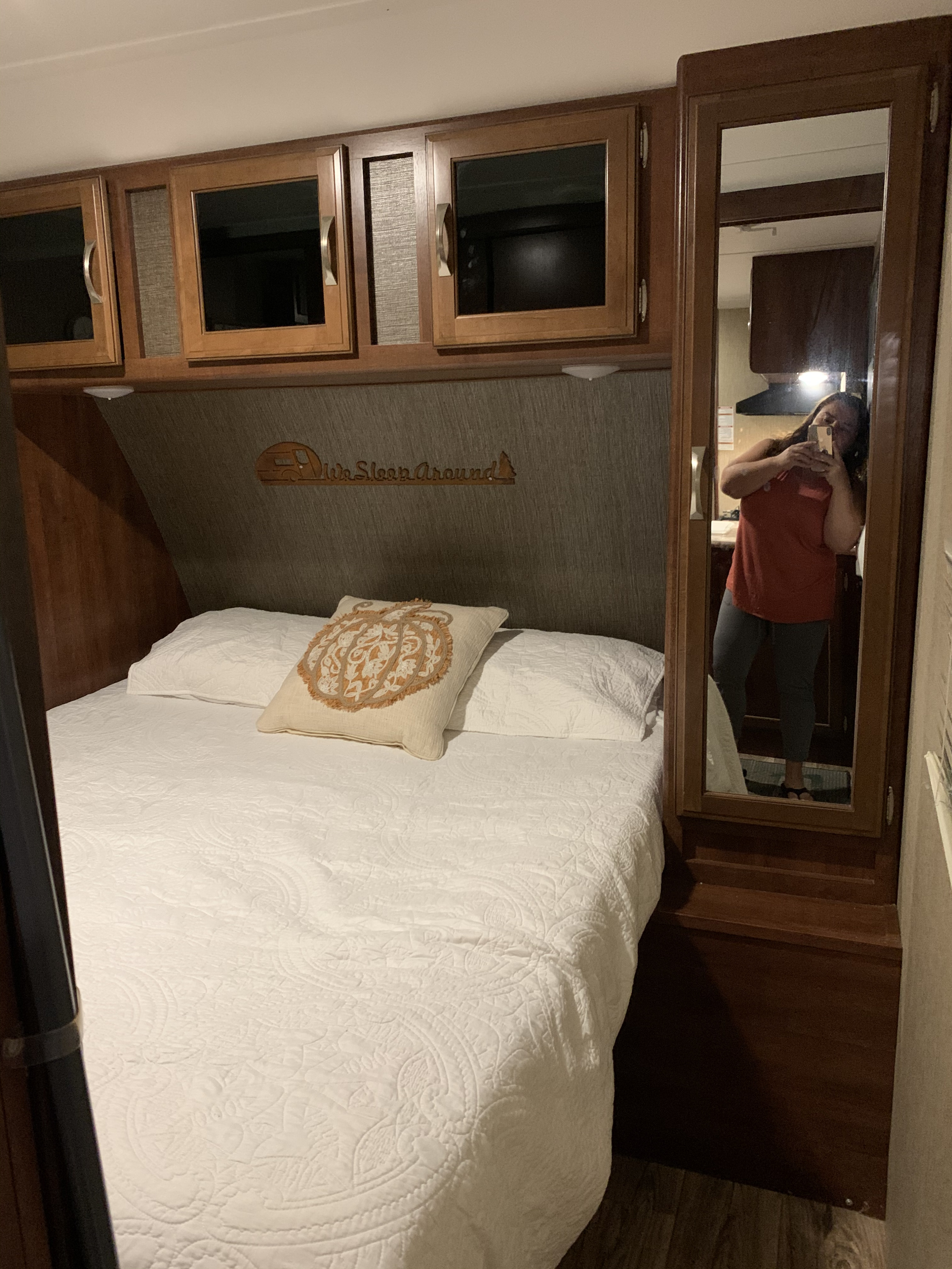 Cozy RV rental bedroom featuring a neatly made bed, overhead storage, and fun "We Sleep Around" decor. User reflected in mirror.. Keystone RV Passport Elite 2017