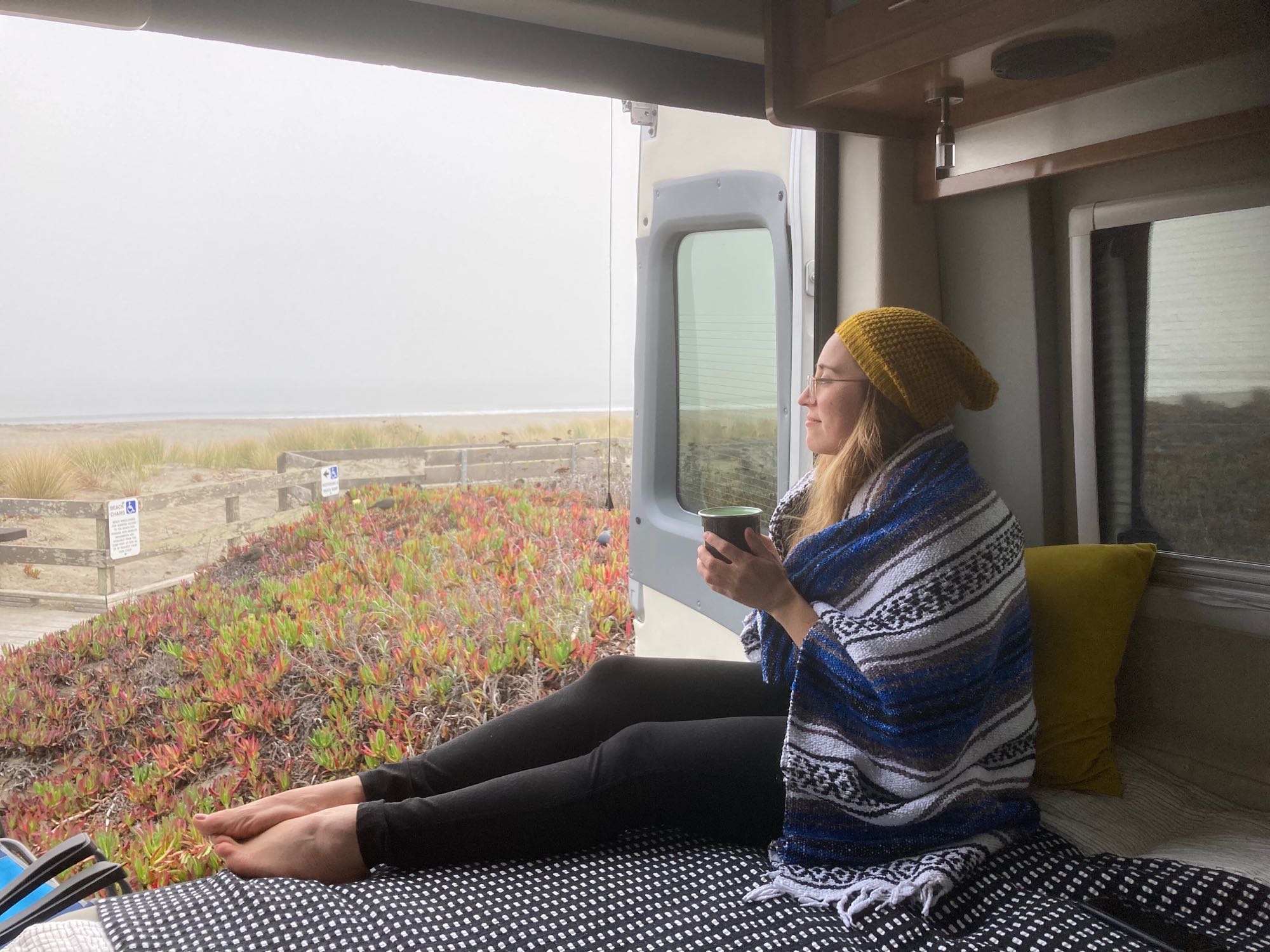 Woman sips coffee from cozy RV rental, enjoying a misty beach view with red and green plants outside.. Ford Other 2020