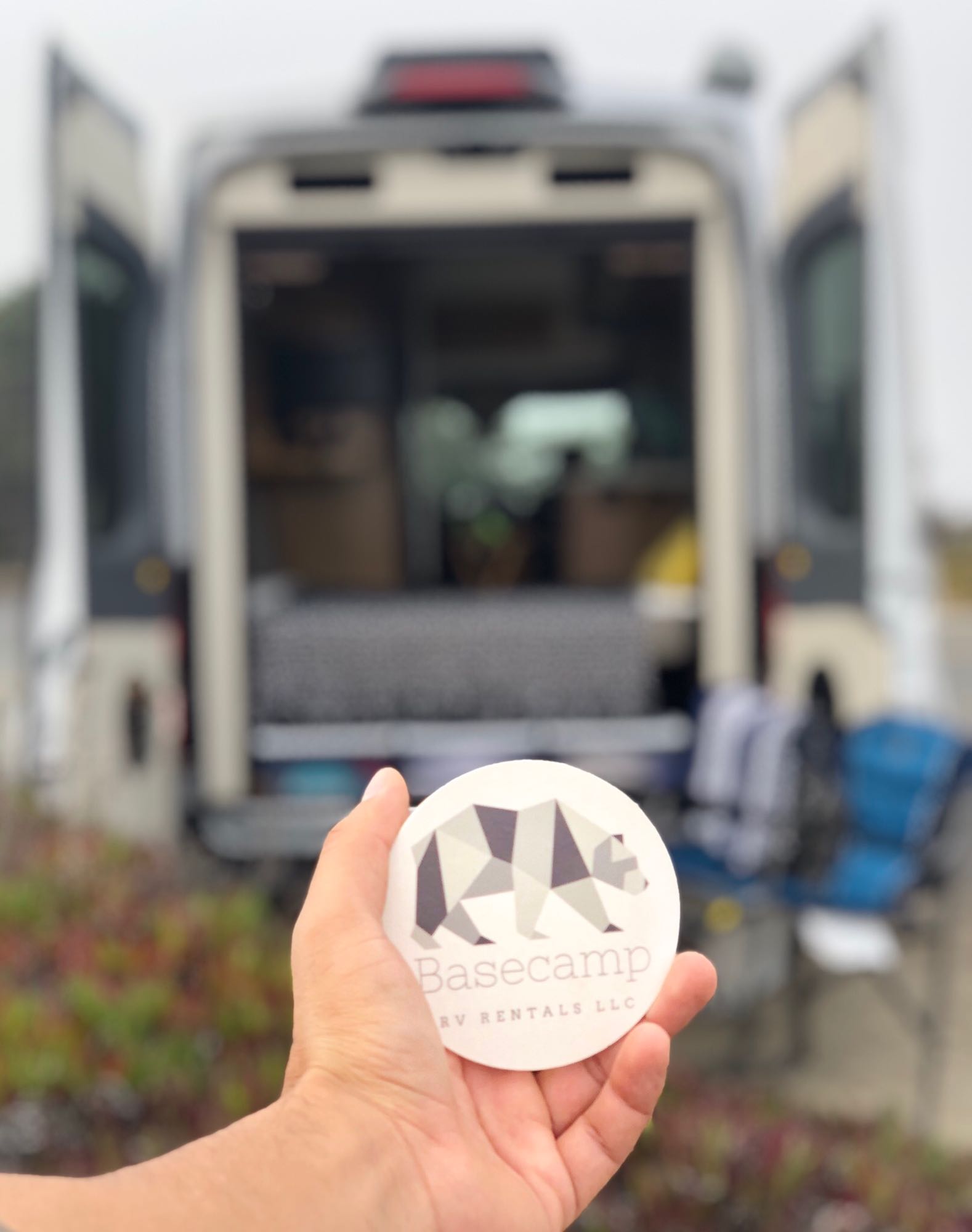 Hand holds a Basecamp RV Rentals logo sticker with a camper van in the blurry background. Perfect for your next adventure!. Ford Other 2020