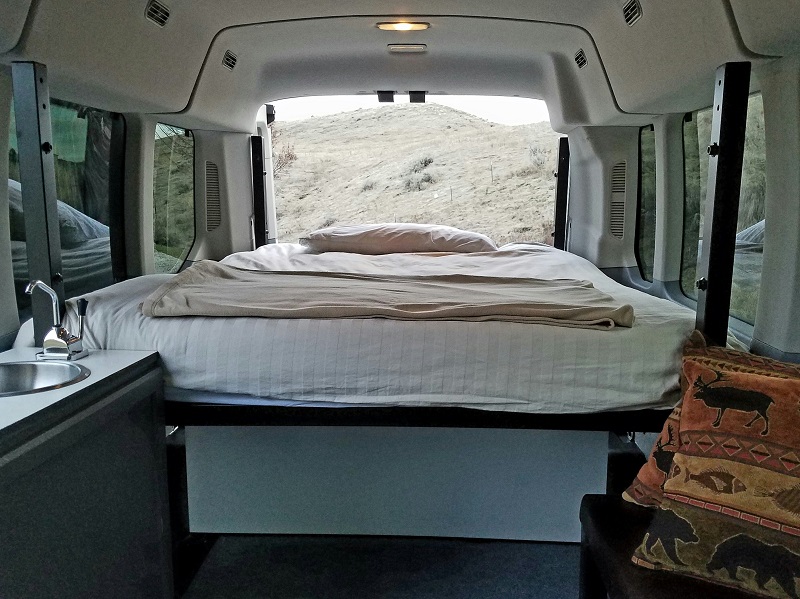 Cozy RV rental interior with a bed, small sink, and open back doors revealing a dry, grassy hill.. Ford transit 2019
