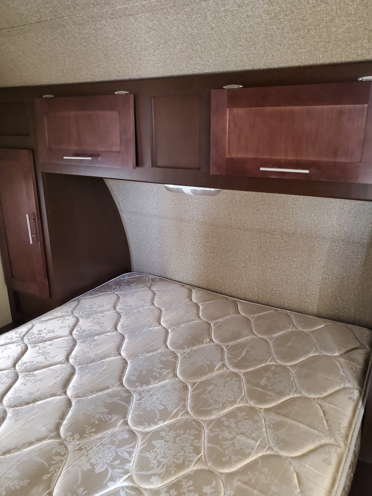 Inside an RV rental: a cozy bedroom with mattress, wood-look overhead cabinets, and upholstered headboard with light.. Forest River Cherokee Grey Wolf 2016