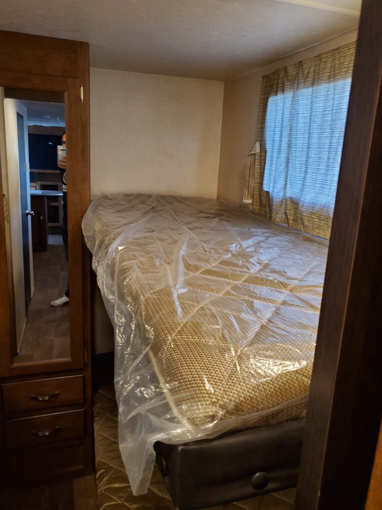 Compact RV rental sleeping area features a bed with a clear plastic cover, wooden cabinet, and window blinds.. Forest River Wildwood X-Lite 2018