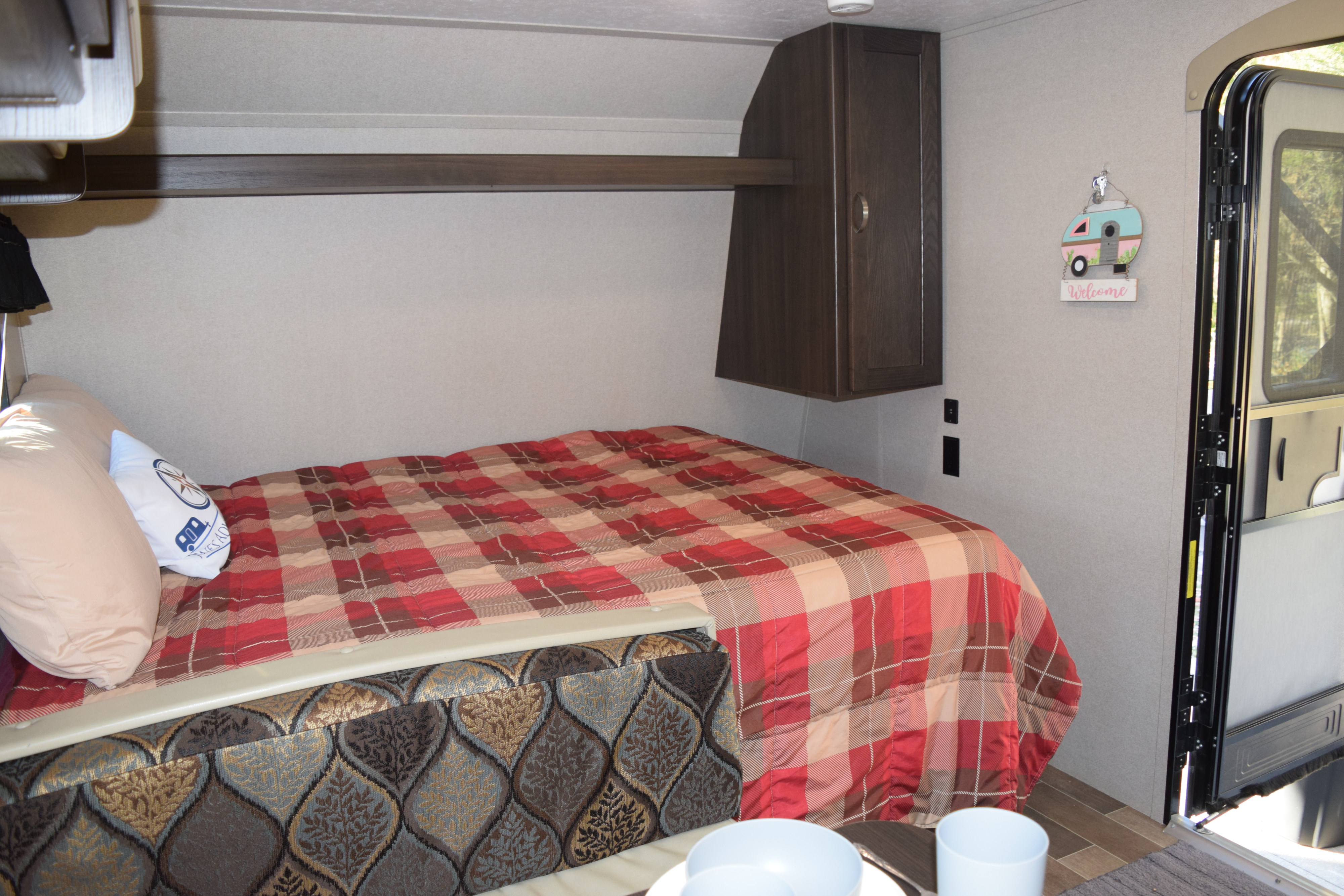 Step inside this RV rental: a cozy sleeping area with plaid bedding, overhead storage, and a 'Welcome' camper sign.. Keystone Springdale 2019