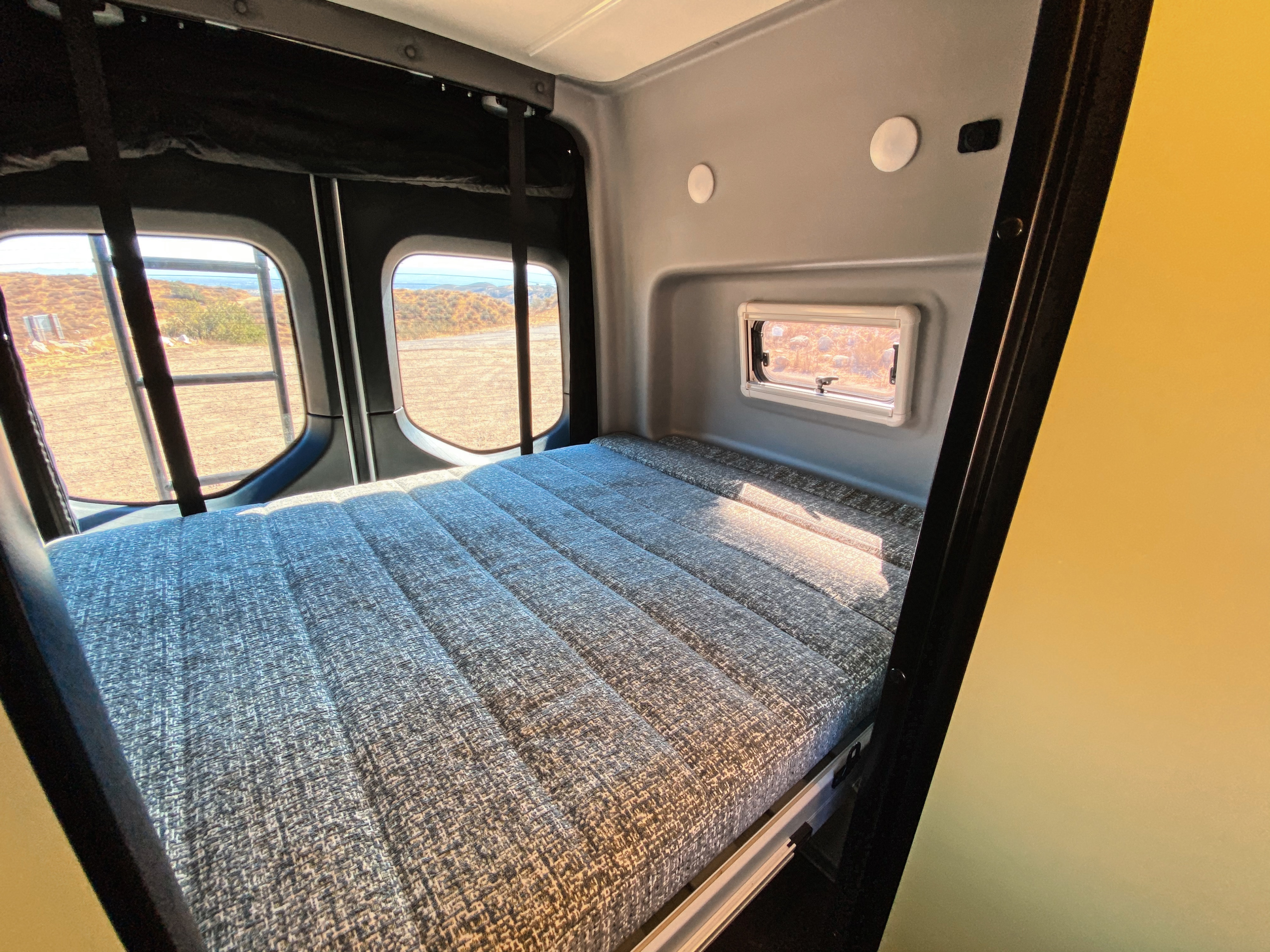 Inside an RV rental: a comfortable bed with a textured gray mattress, flanked by scenic rear windows overlooking a dry landscape.. Winnebago Revel 2020