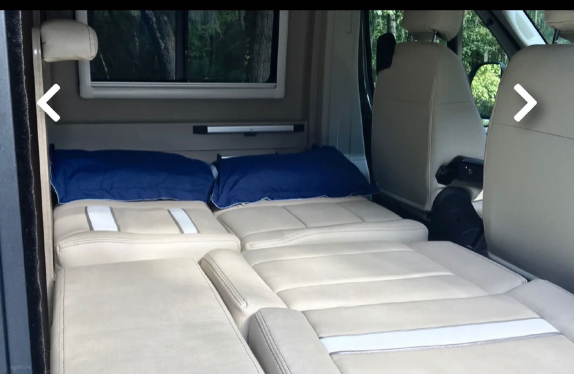 Inside an RV rental: a comfortable beige bed with blue pillows, extending from the rear toward swiveled front seats, ready for travel.. Winnebago Travato 2019