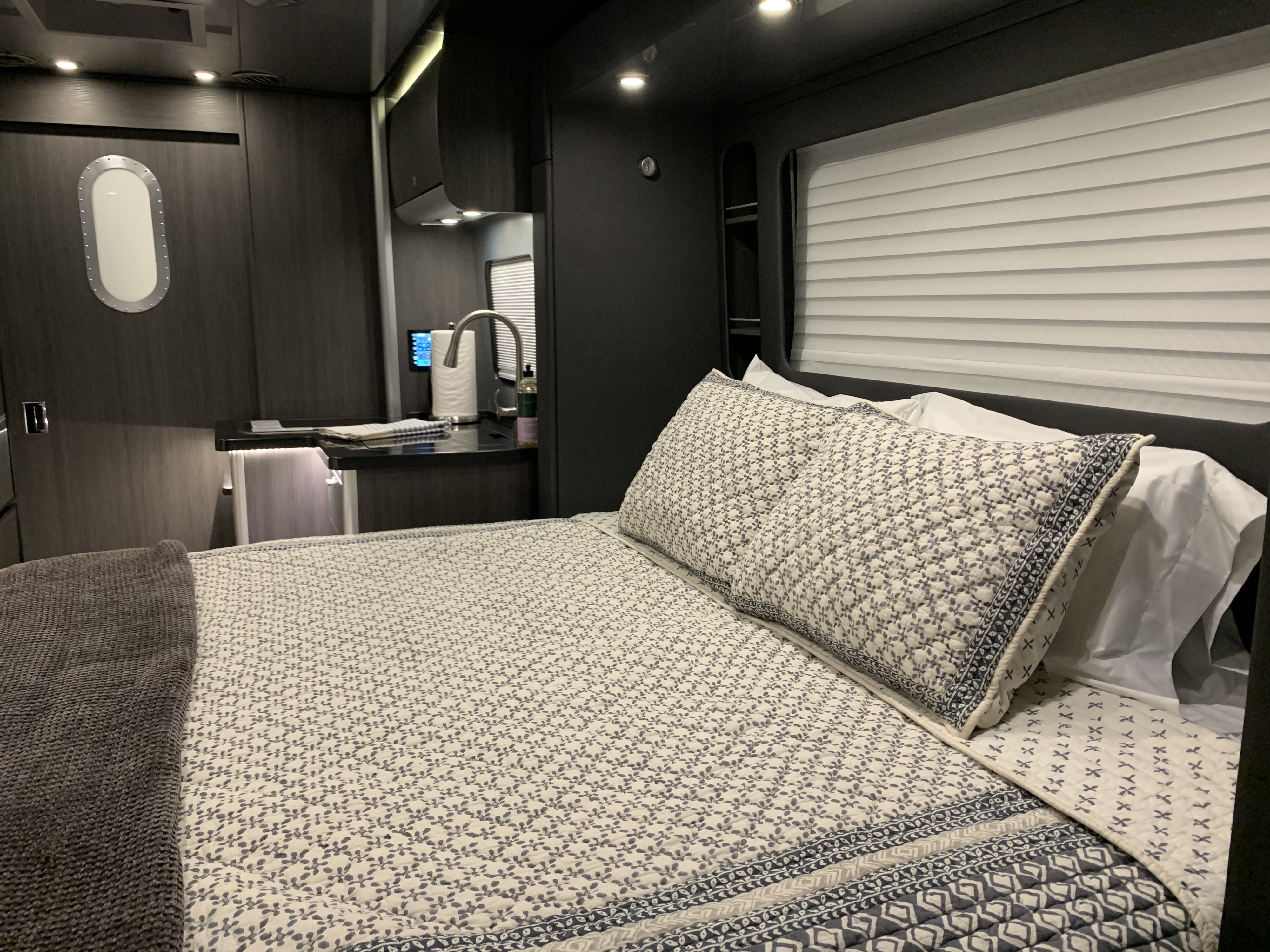 Modern rv rental interior: bedroom with patterned bedding & grey blanket, compact kitchen area, and unique oval window.. Airstream Atlas 2021