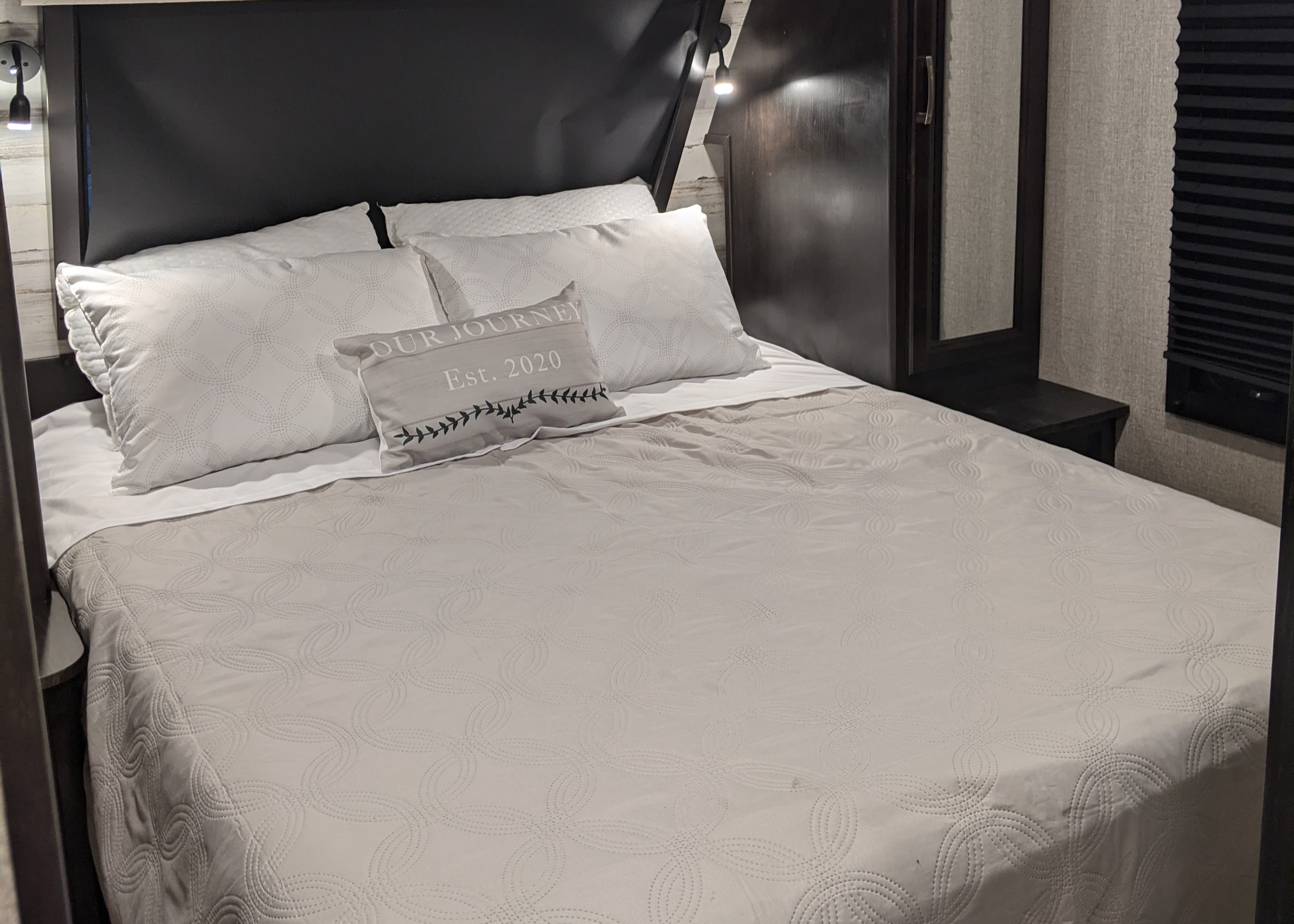 Cozy RV rental bedroom features a comfy bed with quilted bedding, reading lights, and "Our Journey Est. 2020" pillow.. Jayco White Hawk 2021
