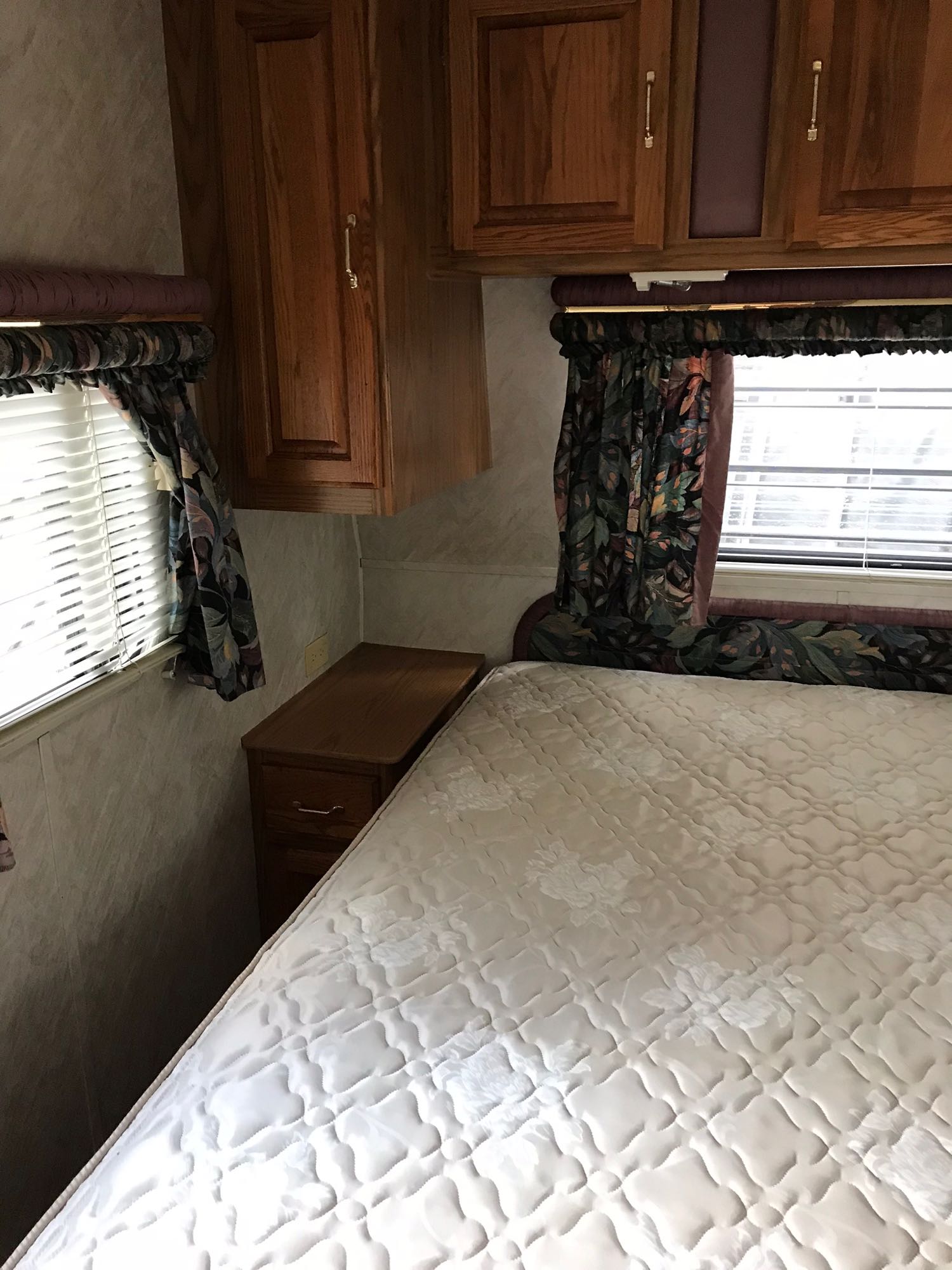 RV rental bedroom: a quilted bed, wooden cabinets, a nightstand, and windows with blinds and leafy patterned curtains.. Jayco Jay Series 1996