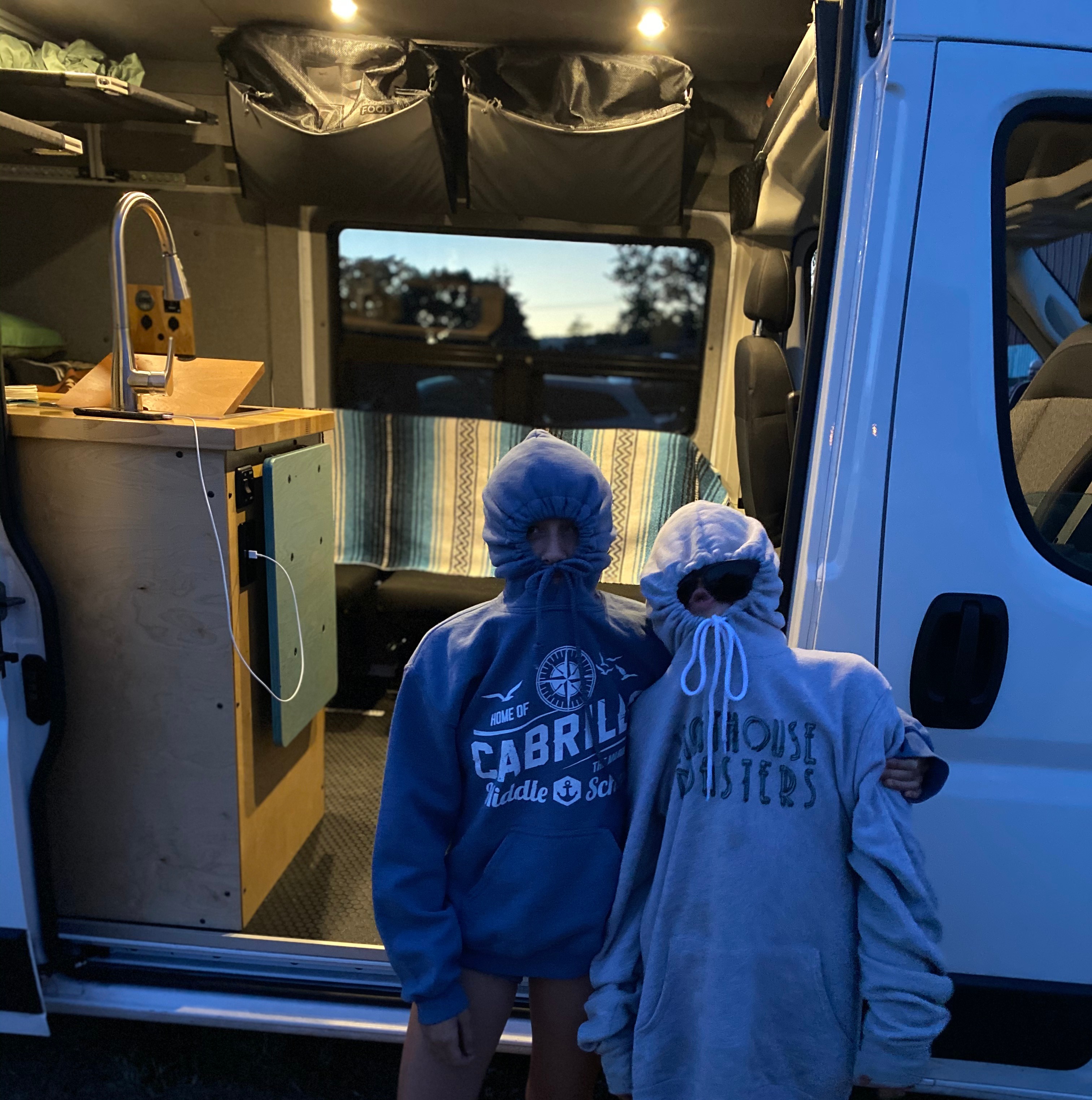 Two people in hoodies stand in an RV rental van, showcasing its cozy interior with a sink, storage, and bench.. Ram Promaster 2500 2019