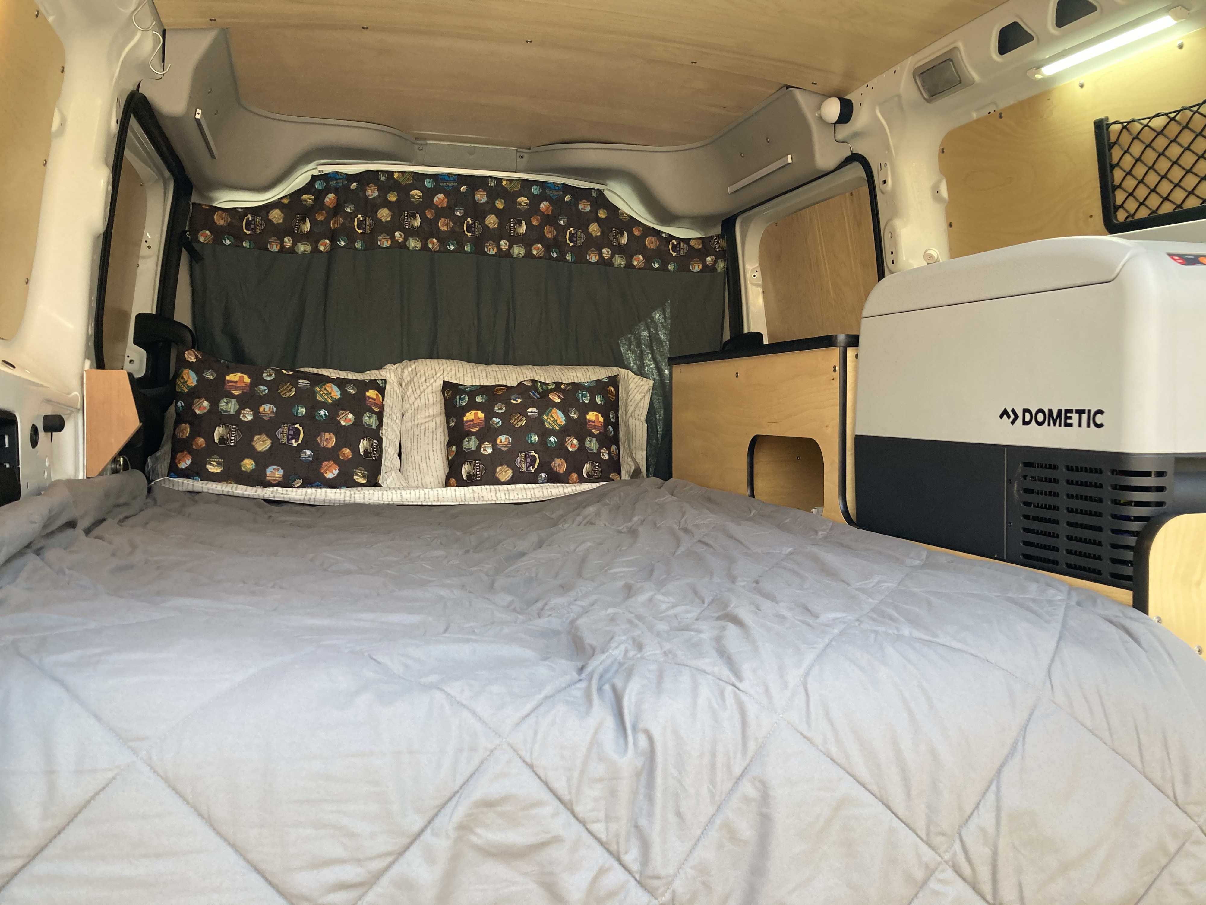 A welcoming camper van bedroom with a soft grey bed, patterned pillows, Dometic fridge, & wood paneling for RV rental.. RAM ProMaster City 2020