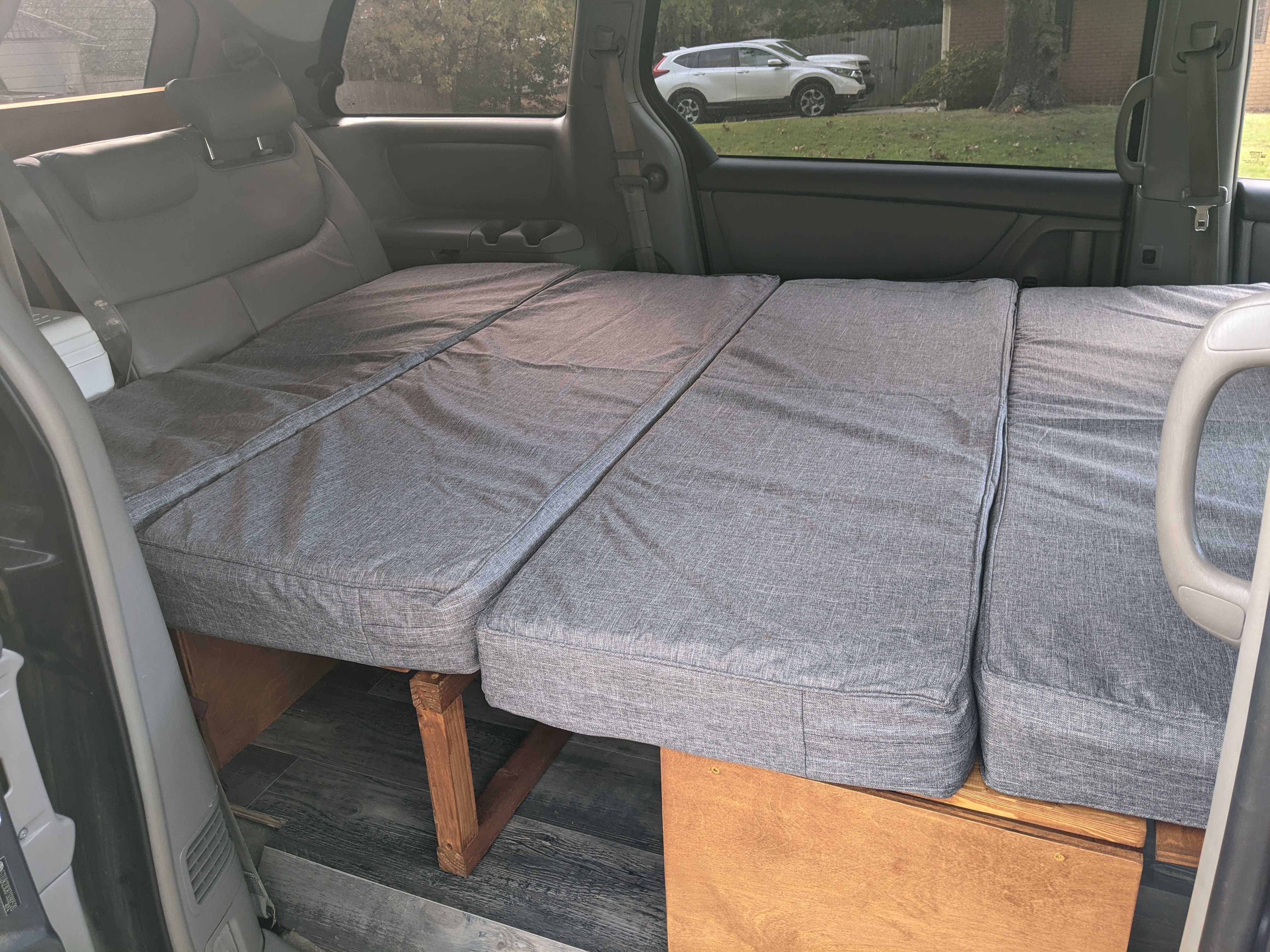 Inside a minivan, a custom-built bed with grey cushions provides a comfy sleeping area, great for an rv rental campervan.. Toyota Other 2007