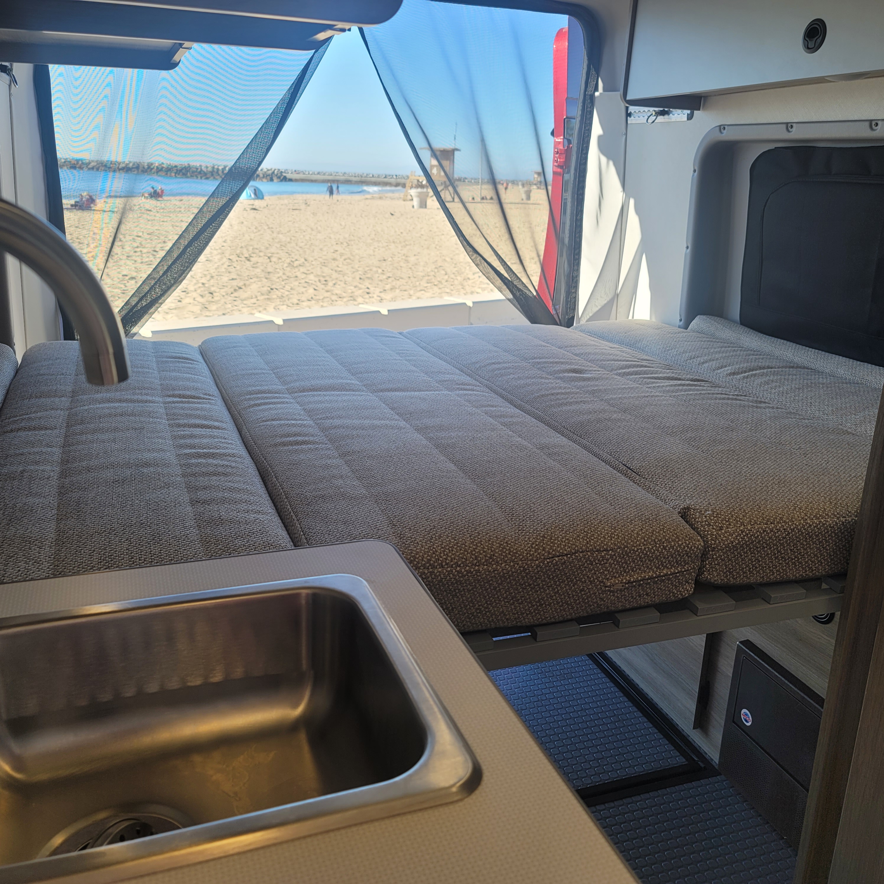RV rental interior with a cozy bed, kitchen sink, and a picturesque beach view through a protective mesh screen.. Winnebago Solis 2021