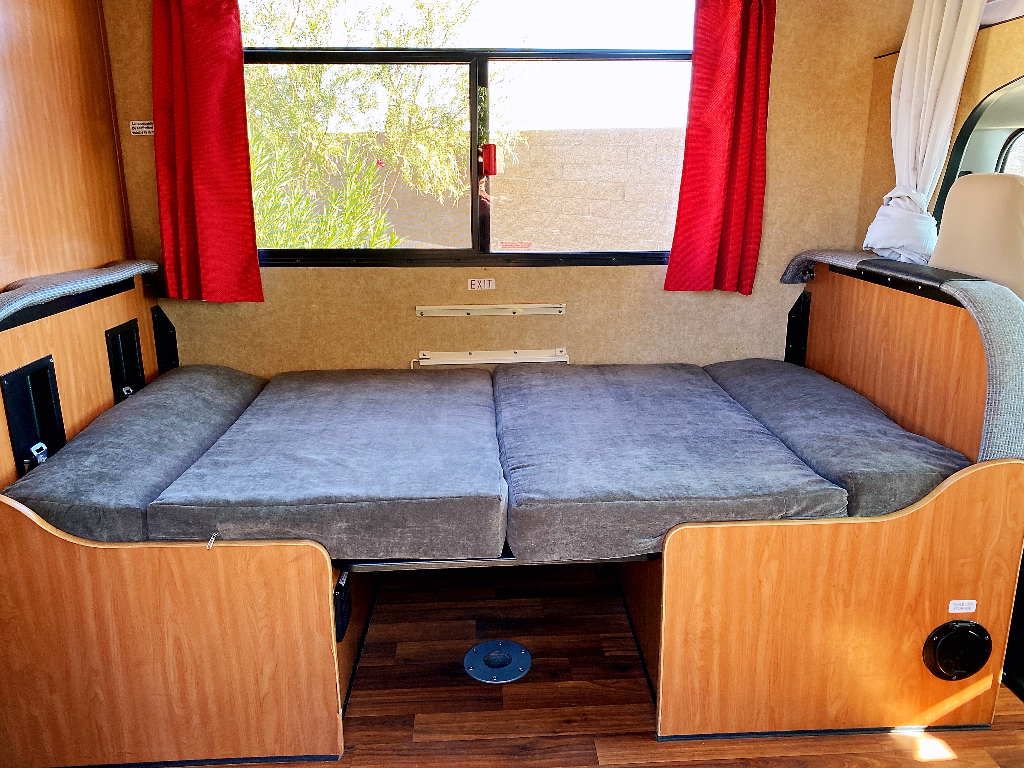 Inside an RV rental: a plush gray bed ready for rest, framed by a wide window with red curtains. A cozy travel setup.. Other Other 2017