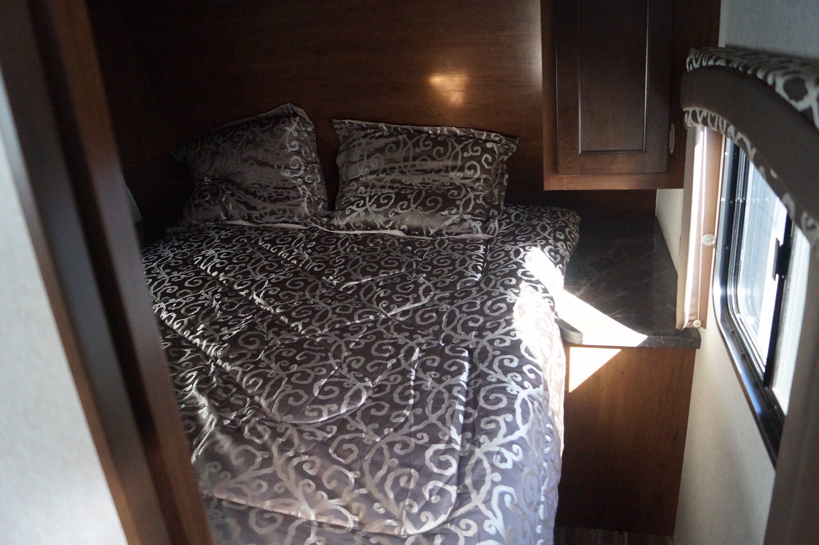 This compact RV rental bedroom features a patterned bed, bedside counter, cabinet, and window, offering a cozy travel space.. Jayco Jay Flight 2017