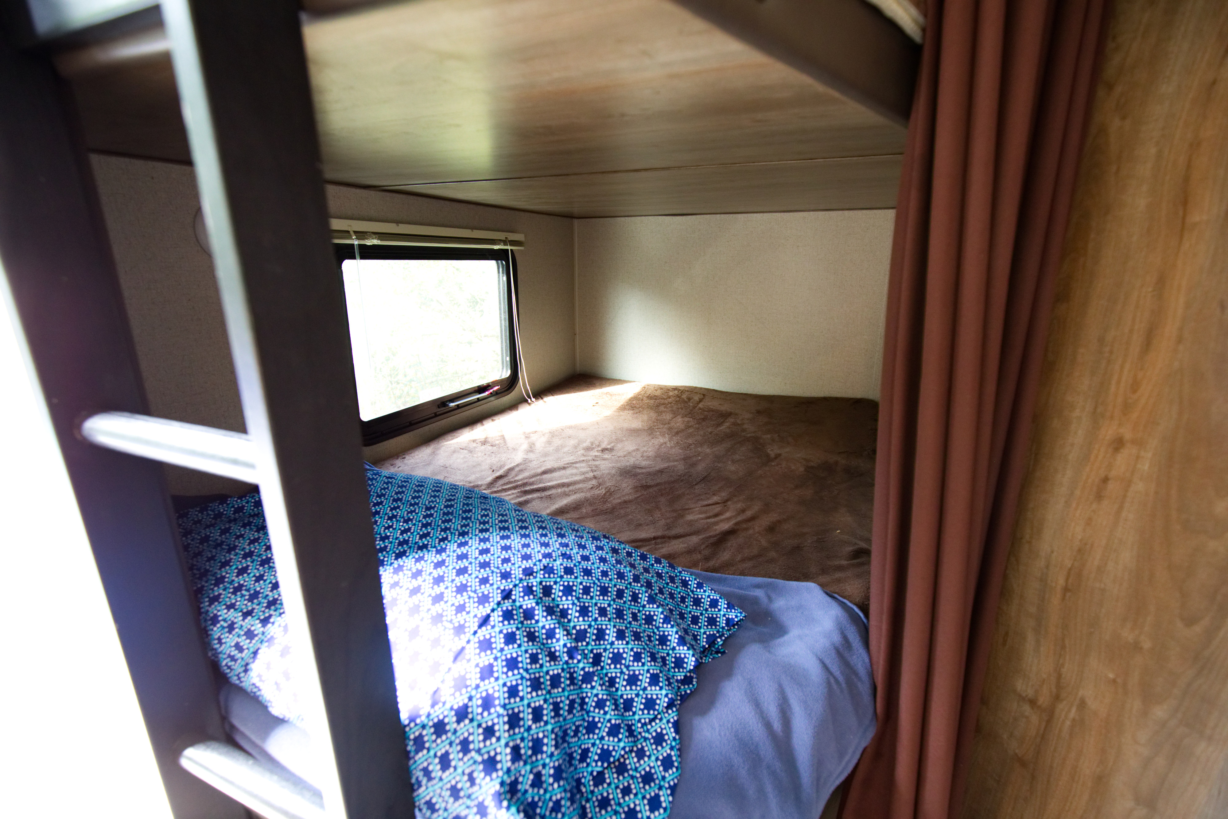 Explore this RV rental's cozy bunk bed, complete with a blue pillow, soft brown blanket, and bright window view.. Other Other 2018